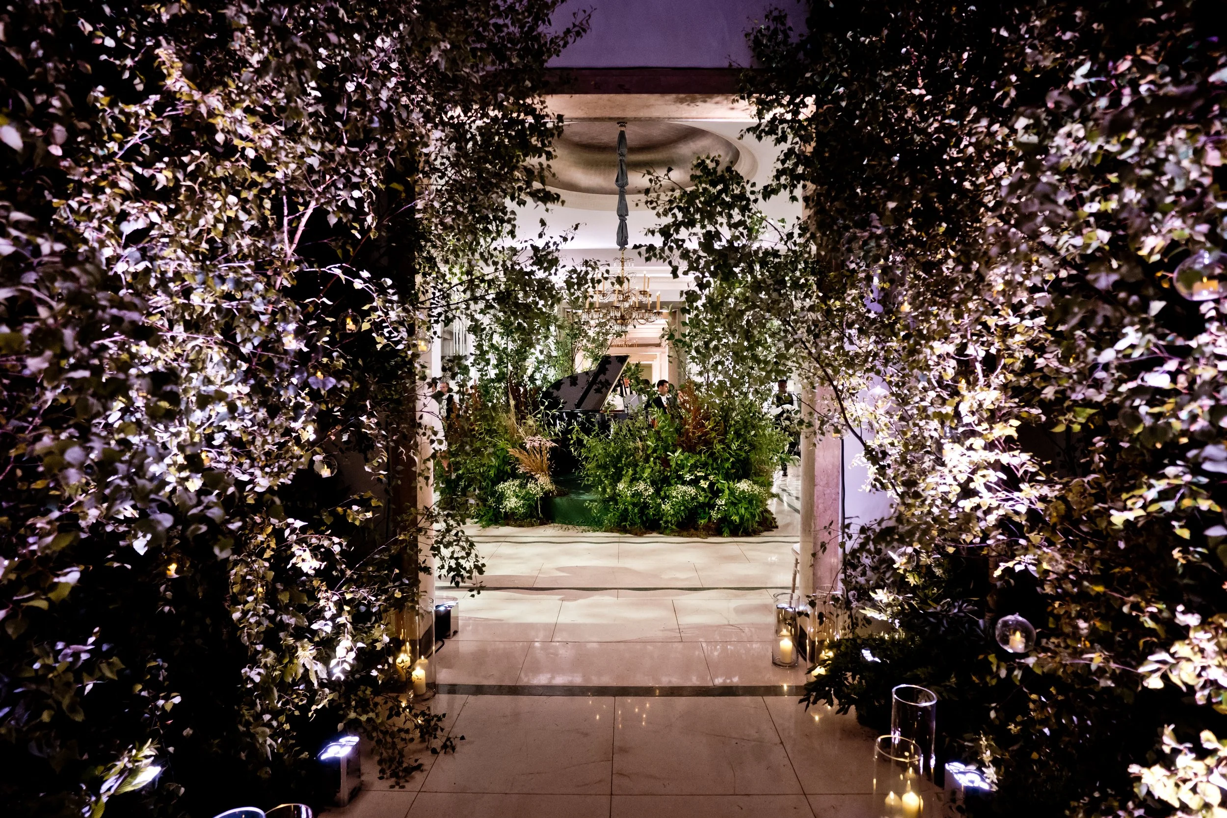 An elegant indoor event space decorated with lush greenery and warm candle lighting, featuring a grand piano and a chandelier, viewed through a framed archway of plants.
