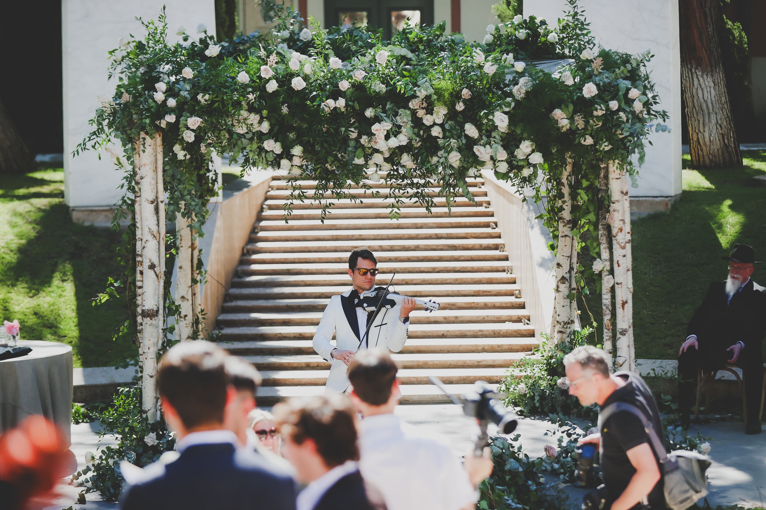 A man in a tuxedo playing the violin at an outdoor wedding ceremony, with guests and floral decorations under a floral-covered archway.