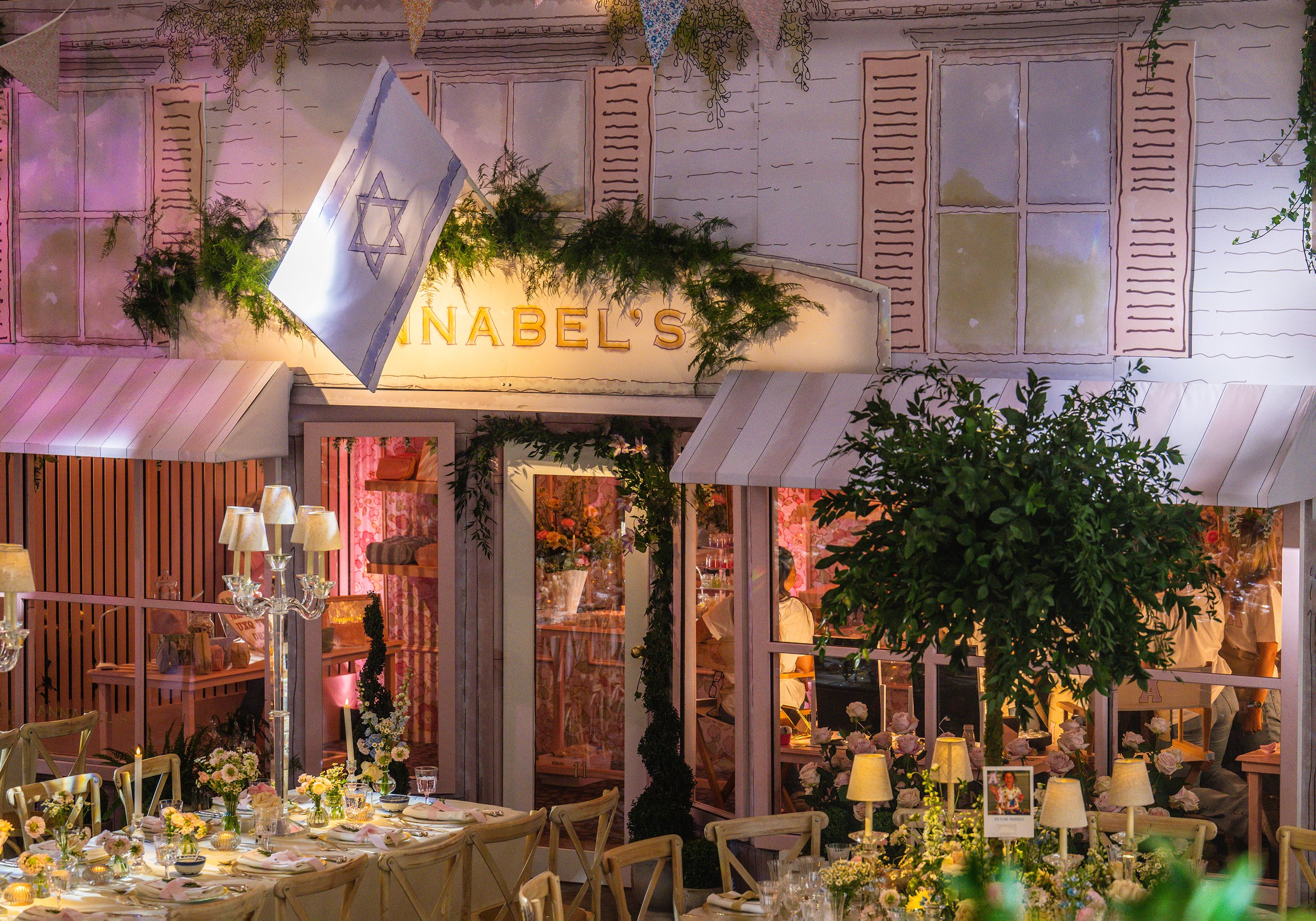 A decorated event space with floral arrangements, elegant table settings, and string lights. A sign with the partially visible word 'NABEL'S' is hung above the windows and greenery is used as decoration.