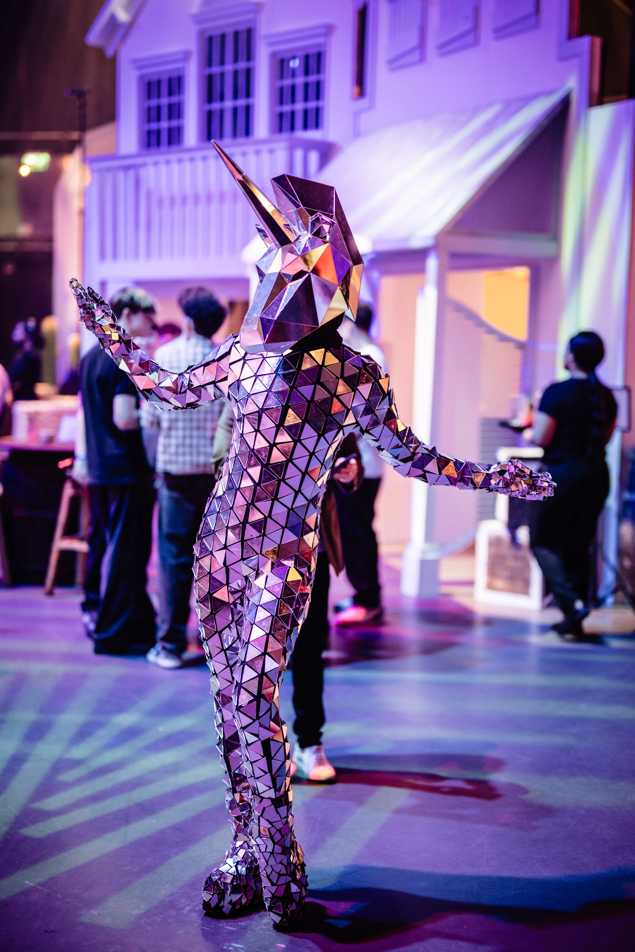 A person dressed in a reflective, geometric unicorn costume with a long horn, dancing at an indoor event with purple and yellow lighting, and a blurred crowd of people in the background at a Bar Mitzvah