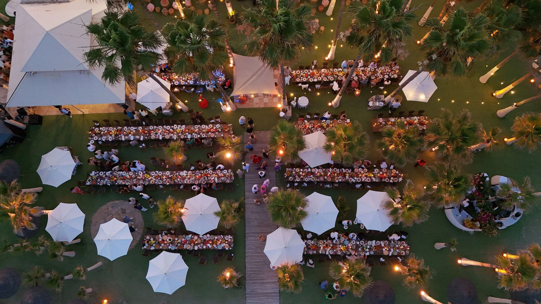 Aerial view of a large outdoor gathering with long banquet tables, white umbrellas, palm trees, and string lights at sunset at Anantara Villa Padierna Resort in Marbella