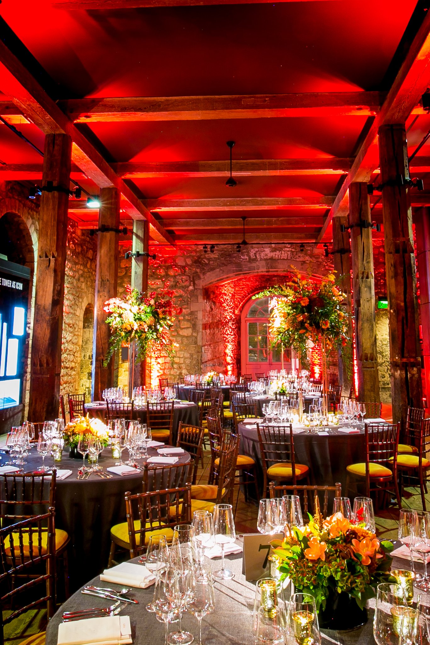 Set dining tables with floral centerpieces, glassware, candles, and place settings in an elegant event space with wooden beams, stone walls, and red lighting at the Tower of London