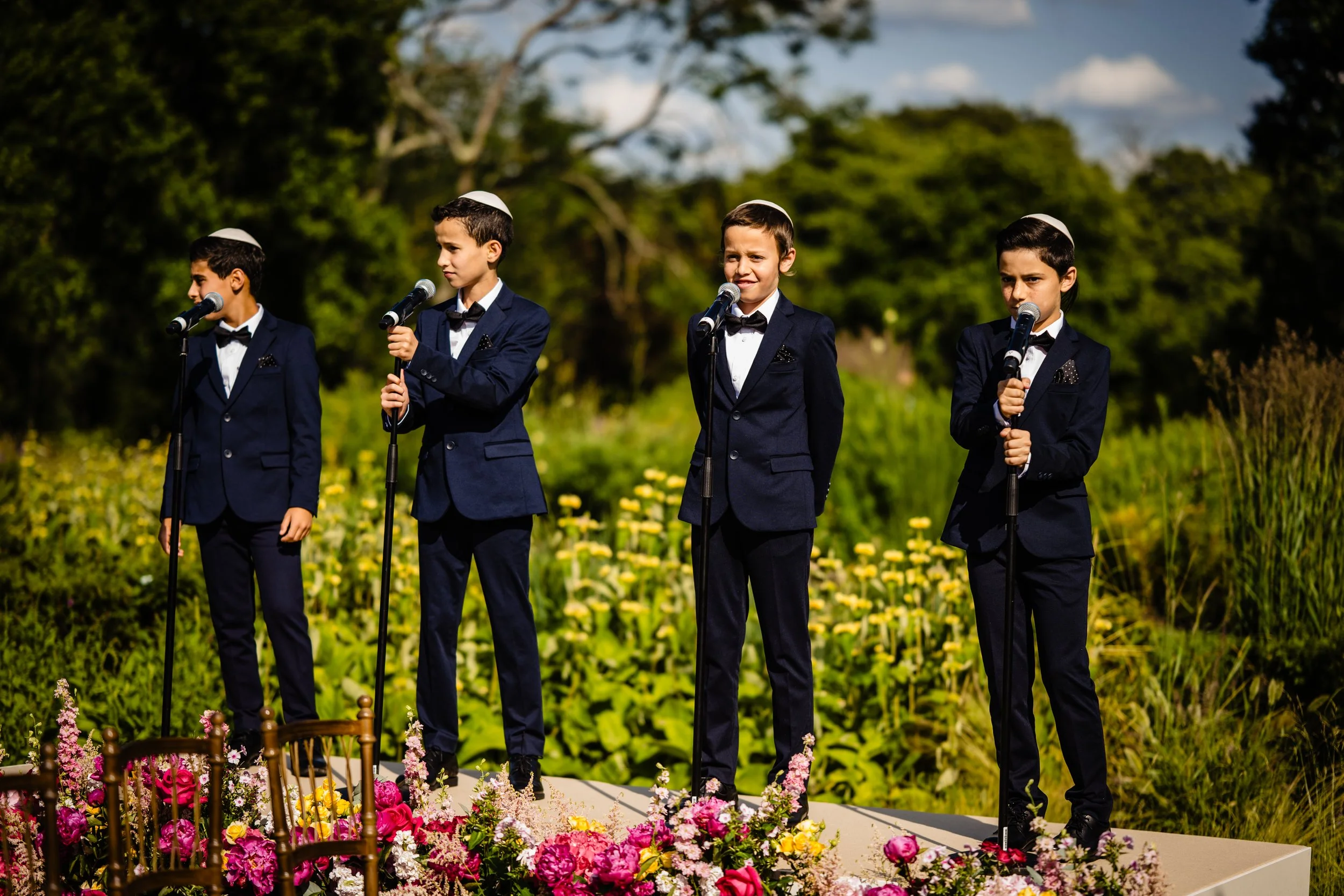 Four boys dressed in formal navy suits with black bow ties, holding microphones and wearing white kippahs, stand outdoors on a stage decorated with pink and red flowers, with a lush green background. Fairmont Windsor Park