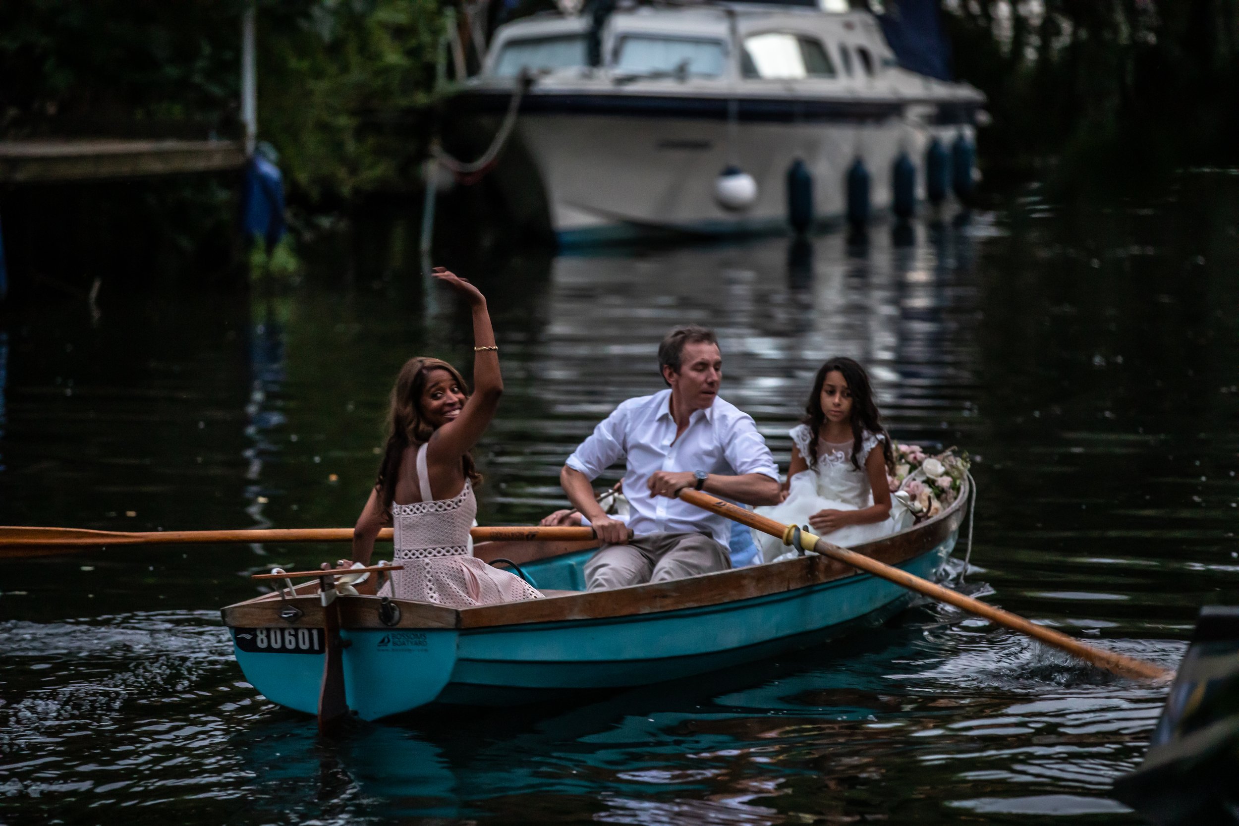 Three people are in a small boat on a calm river. One woman is smiling and waving, another man is rowing, and a young girl is sitting next to him, holding flowers. There are boats docked along the riverbank in the background.