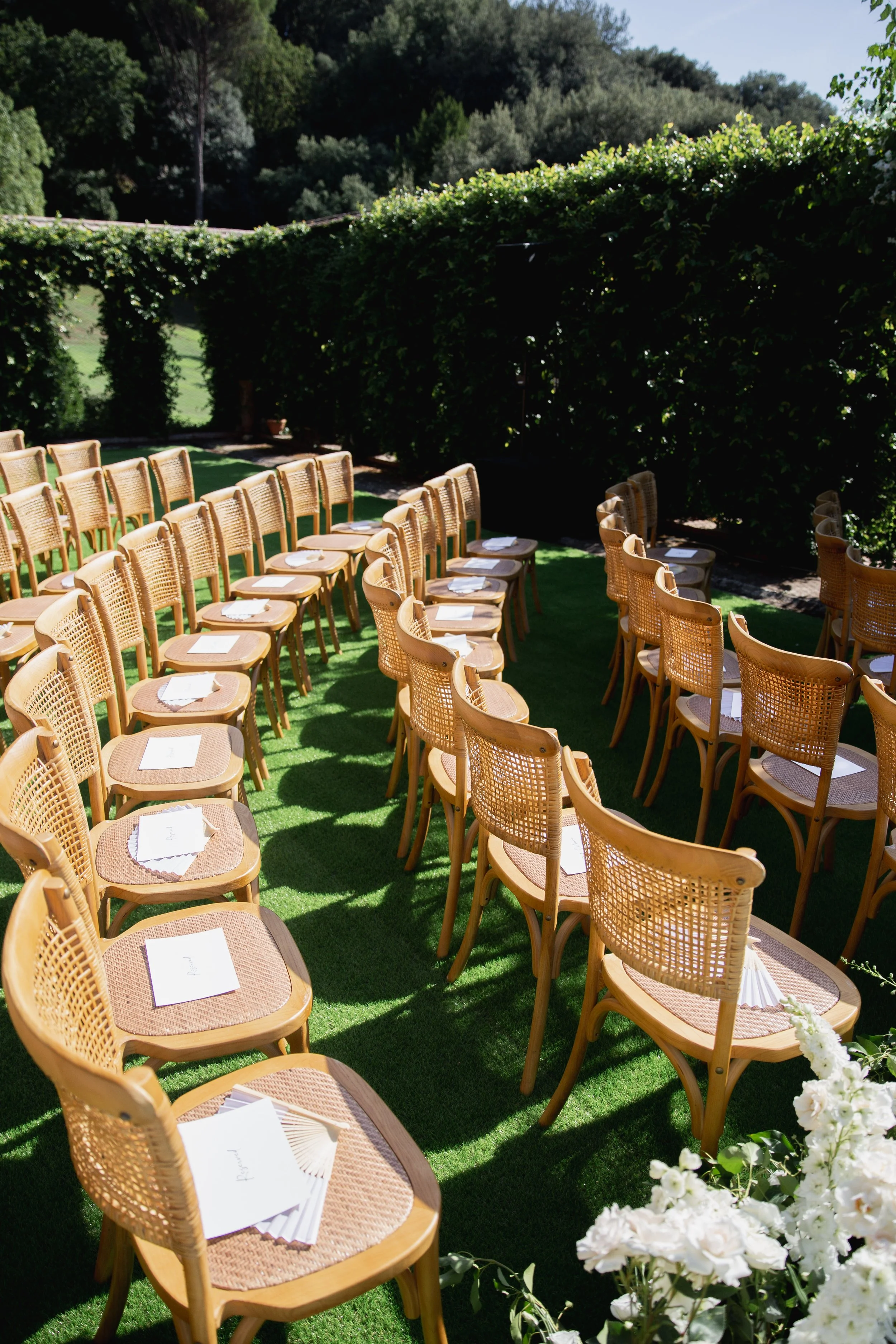 Outdoor seating area with multiple tan chairs arranged in rows on green turf, set for an event, with white paper insets on each chair and white flowers decor in the foreground. Green hedges and trees in the background under a clear sky at Villa Palmi