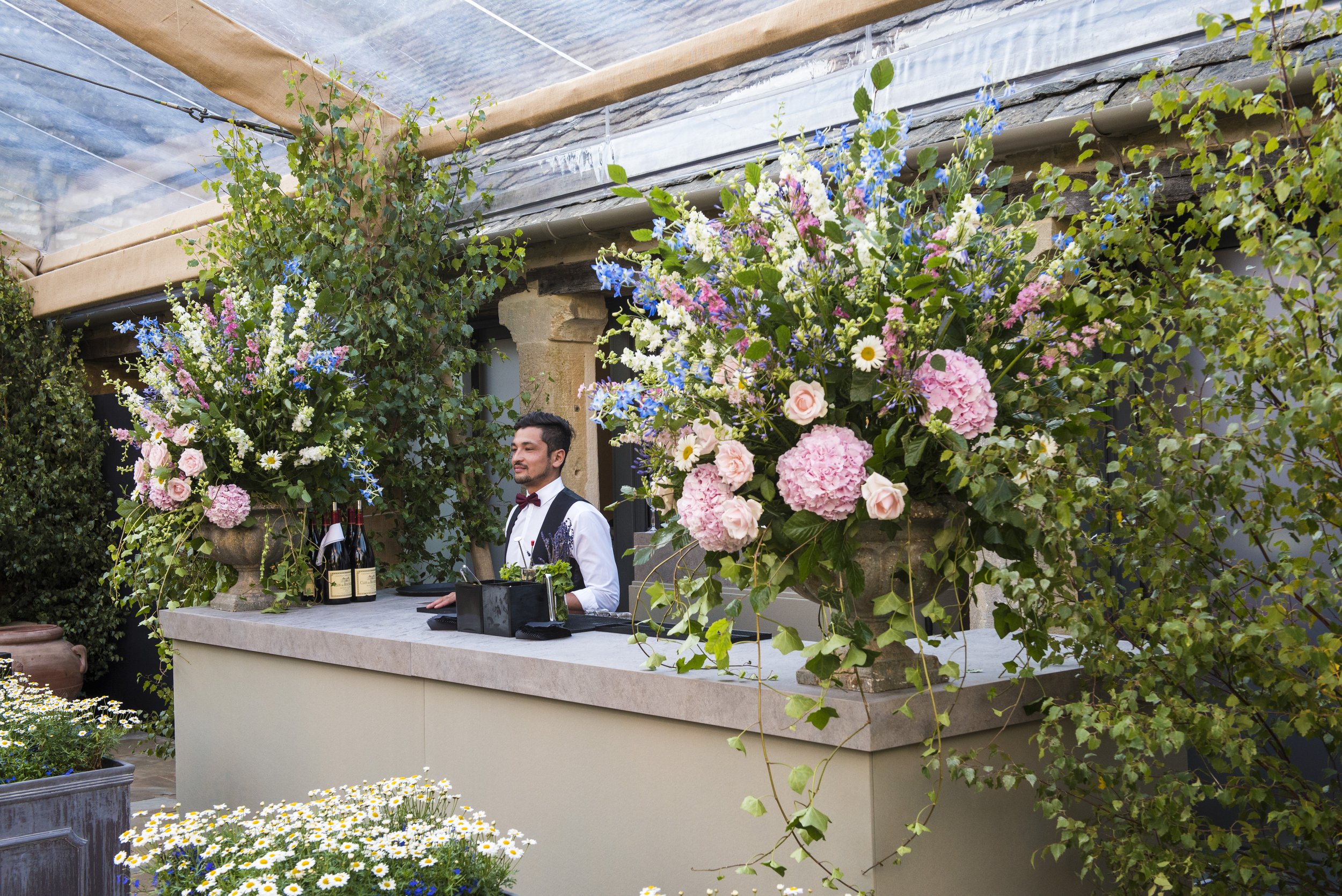 A bartender standing behind an outdoor bar decorated with large flower arrangements, with bottles of wine and a tray on the bar counter, surrounded by greenery and flowers.
