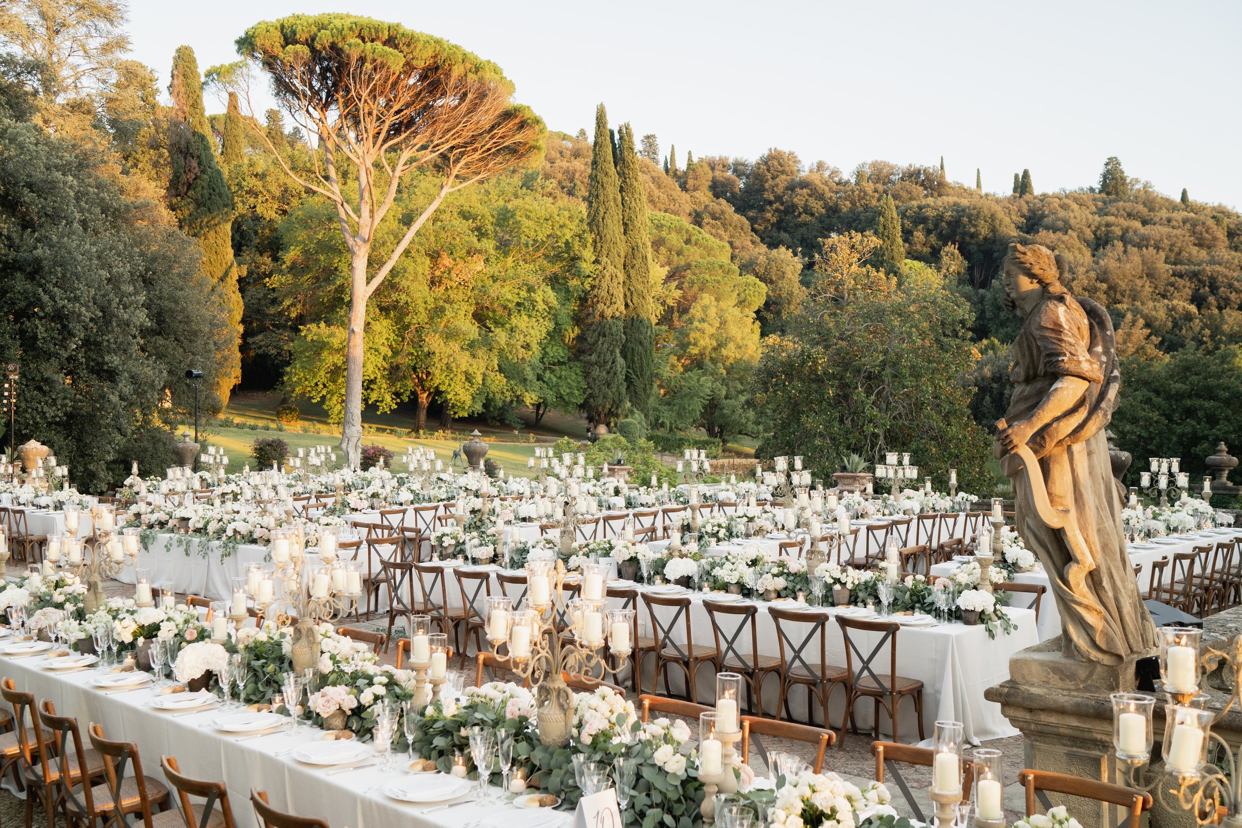 Outdoor wedding reception setup with long tables decorated with white flowers and candles, surrounded by chairs, with a backdrop of lush green trees and a statue in the foreground at Villa Palmieri in Florence