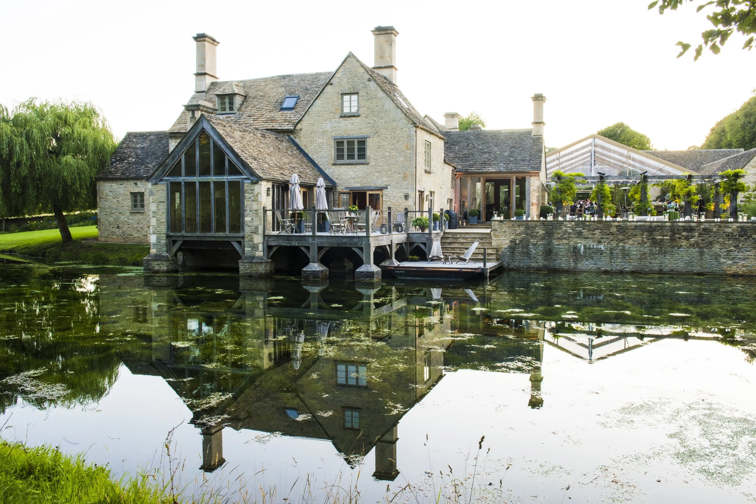 A stone house with a sloped roof and multiple chimneys is reflected in a calm body of water. There is a deck with outdoor furniture and umbrellas extending over the water, surrounded by greenery and trees.