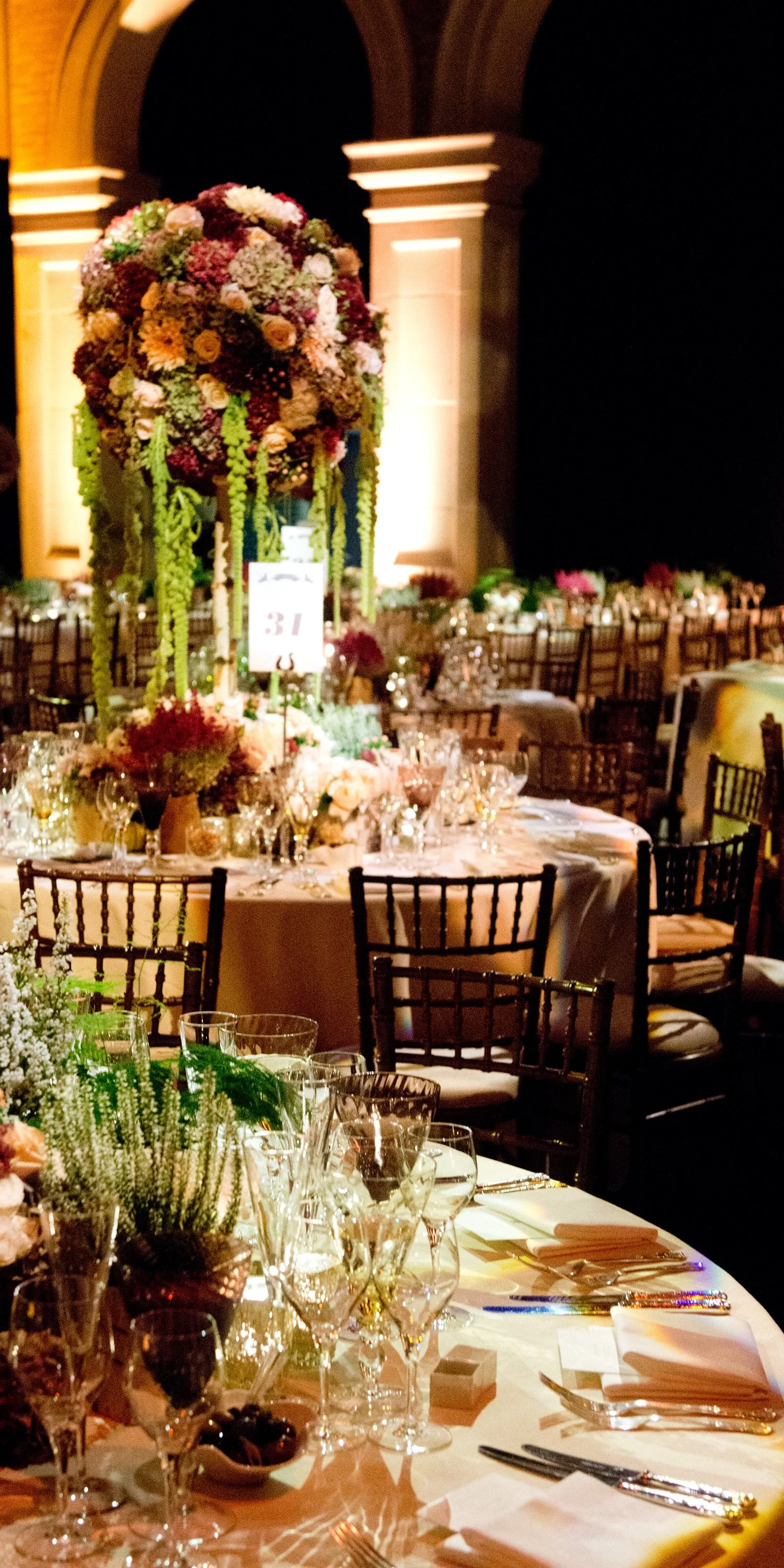 Elegant banquet table setup with numerous glasses, plates, silverware, and a floral centerpiece with cascading flowers, at a formal event in a dimly lit venue.