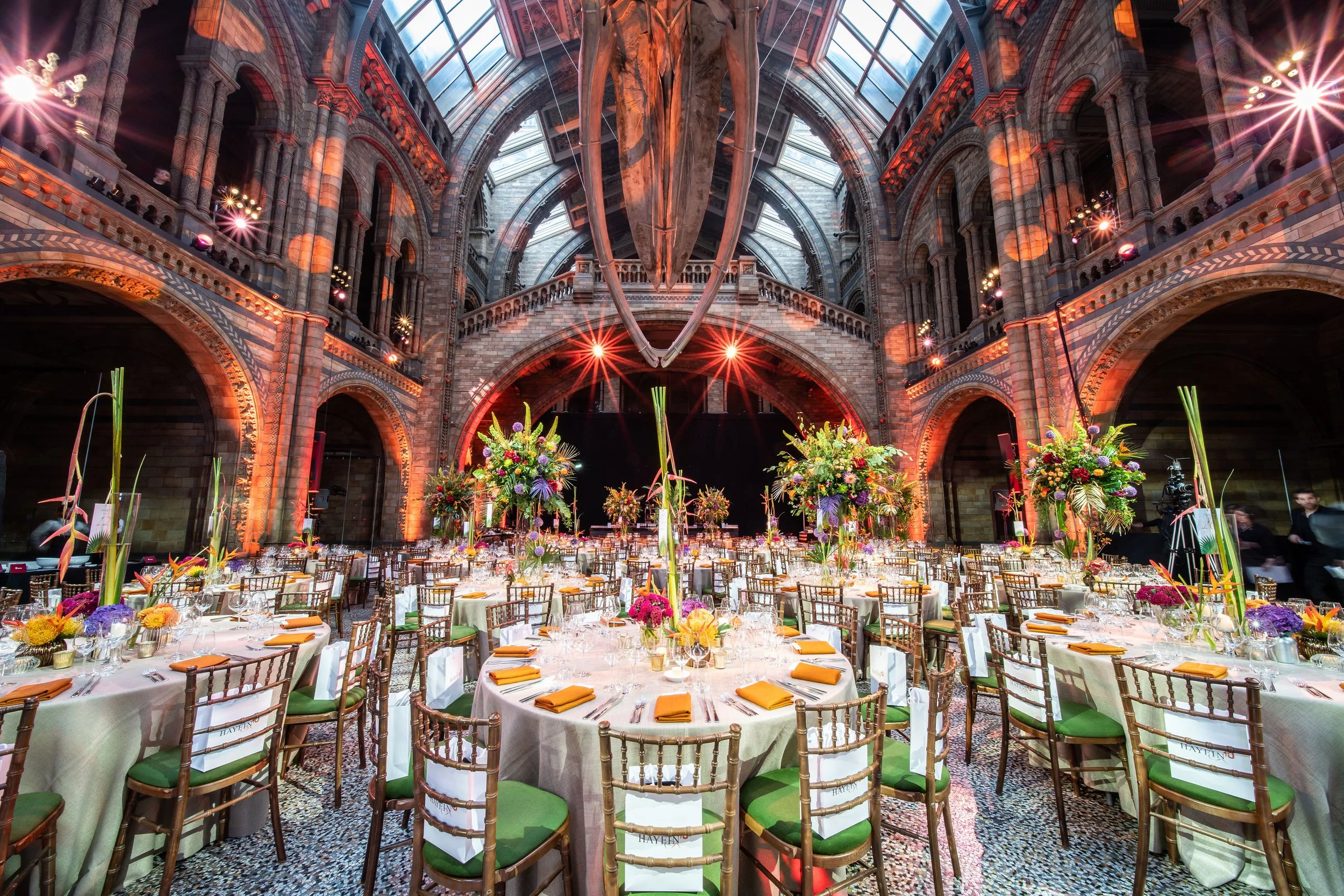 Elegant banquet hall with high arched ceilings and glass skylights, decorated with vibrant floral centerpieces on round tables and rows of chairs, prepared for a corporate event at The Natural History Museum
