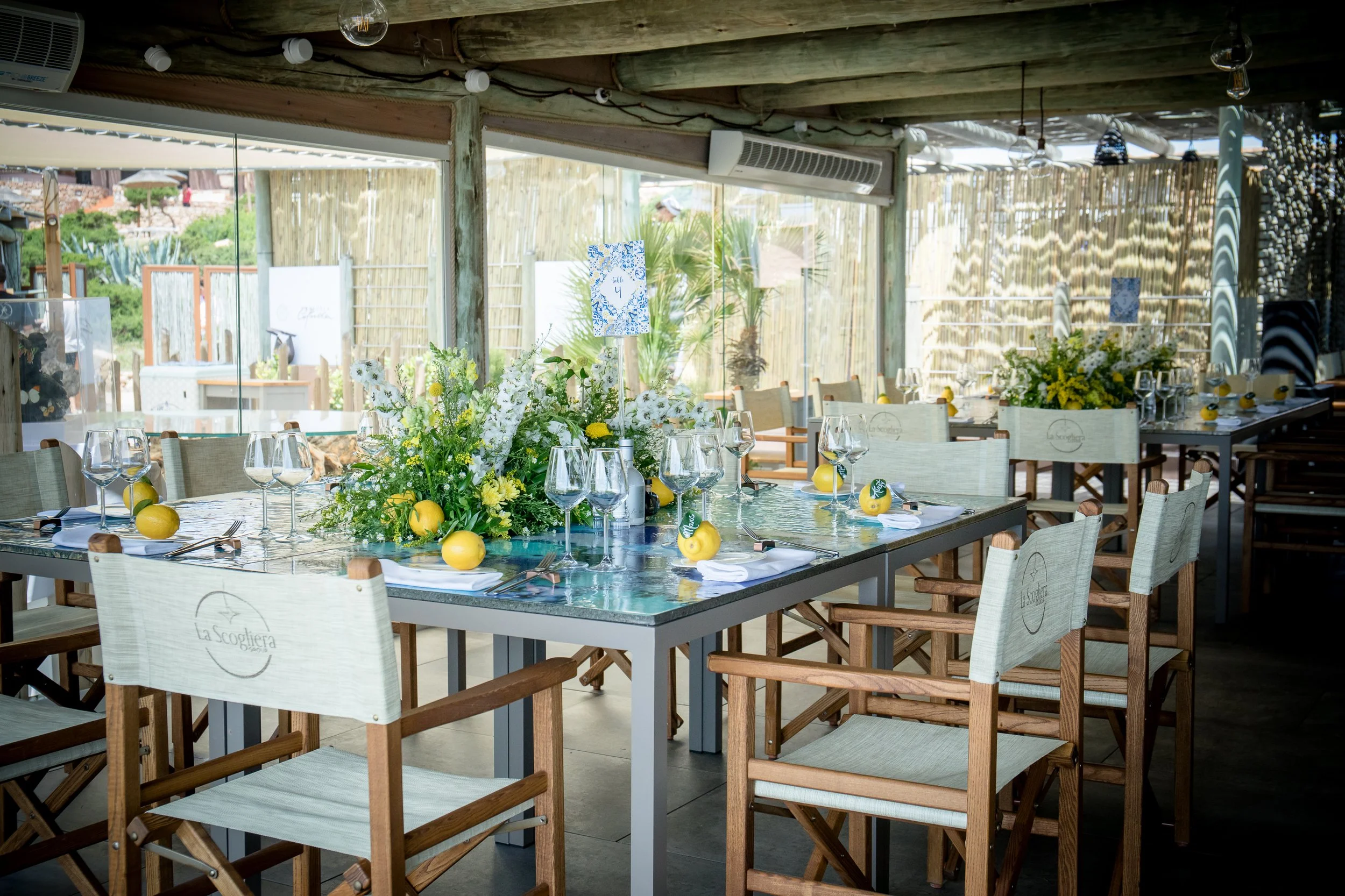 Elegant indoor dining setup with a large rectangular table decorated with white and yellow floral centerpieces, lemons, and wine glasses. The chairs are wooden with beige cushions, and the environment is bright with natural light coming through large