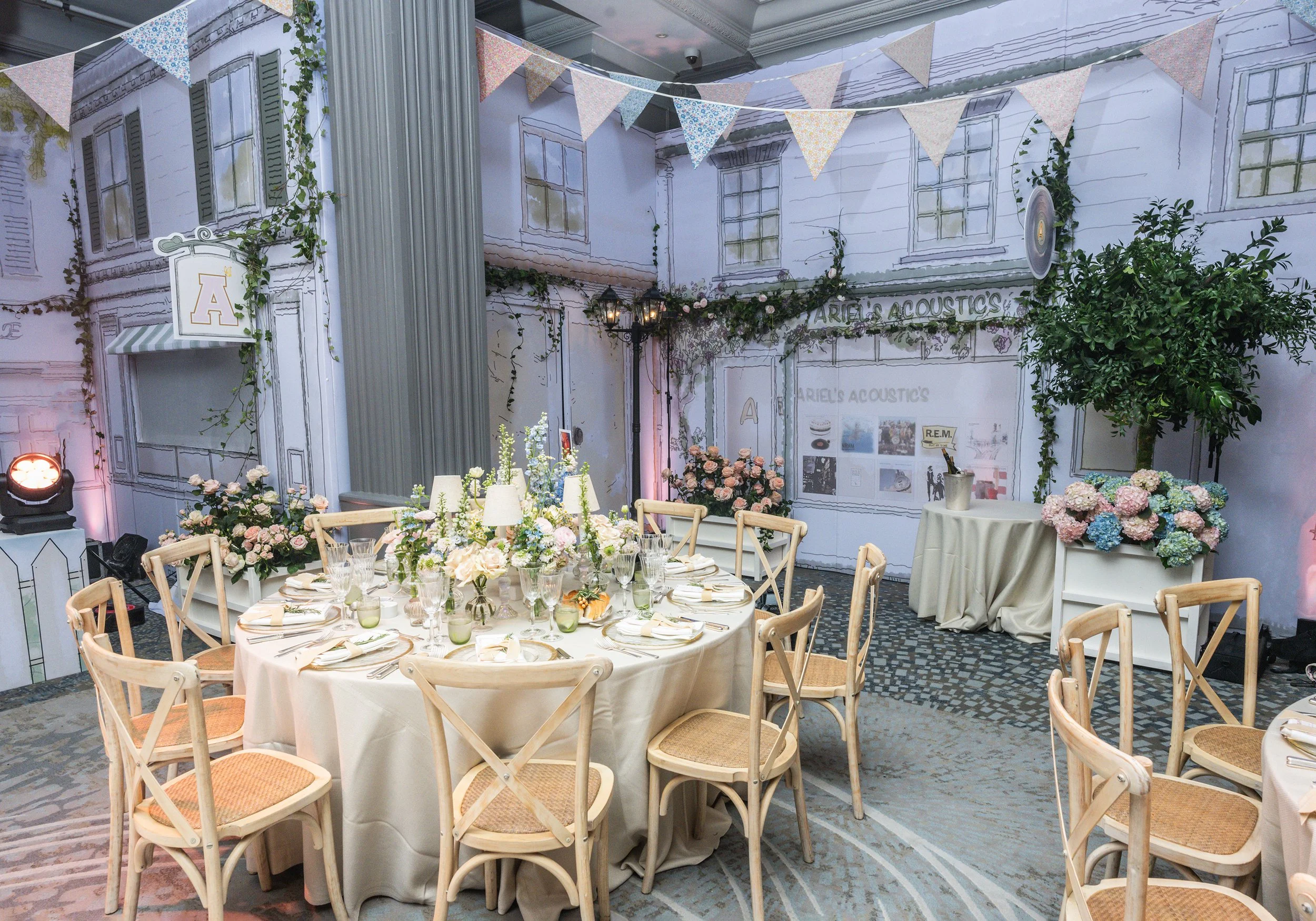 A decorated indoor event space with round tables covered in white tablecloths, set with glassware and napkins, surrounded by wooden chairs with woven seats, and decorated with flowers and greenery. The background features a painted mural of a buildin