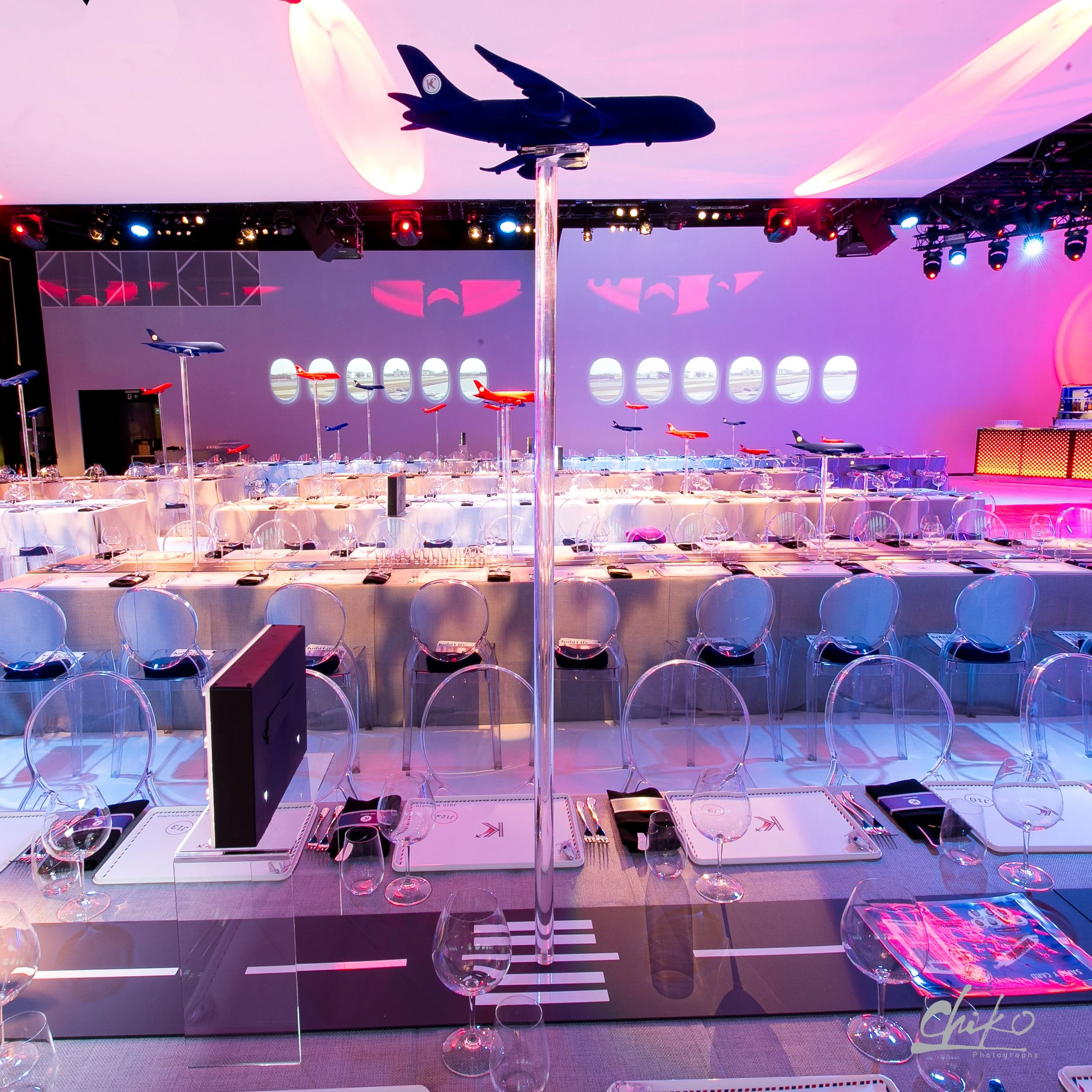 Table set with chairs and tableware, decorated with model airplanes suspended on clear stands, in a brightly lit event space with purple and pink lighting and airplane window designs on the wall.