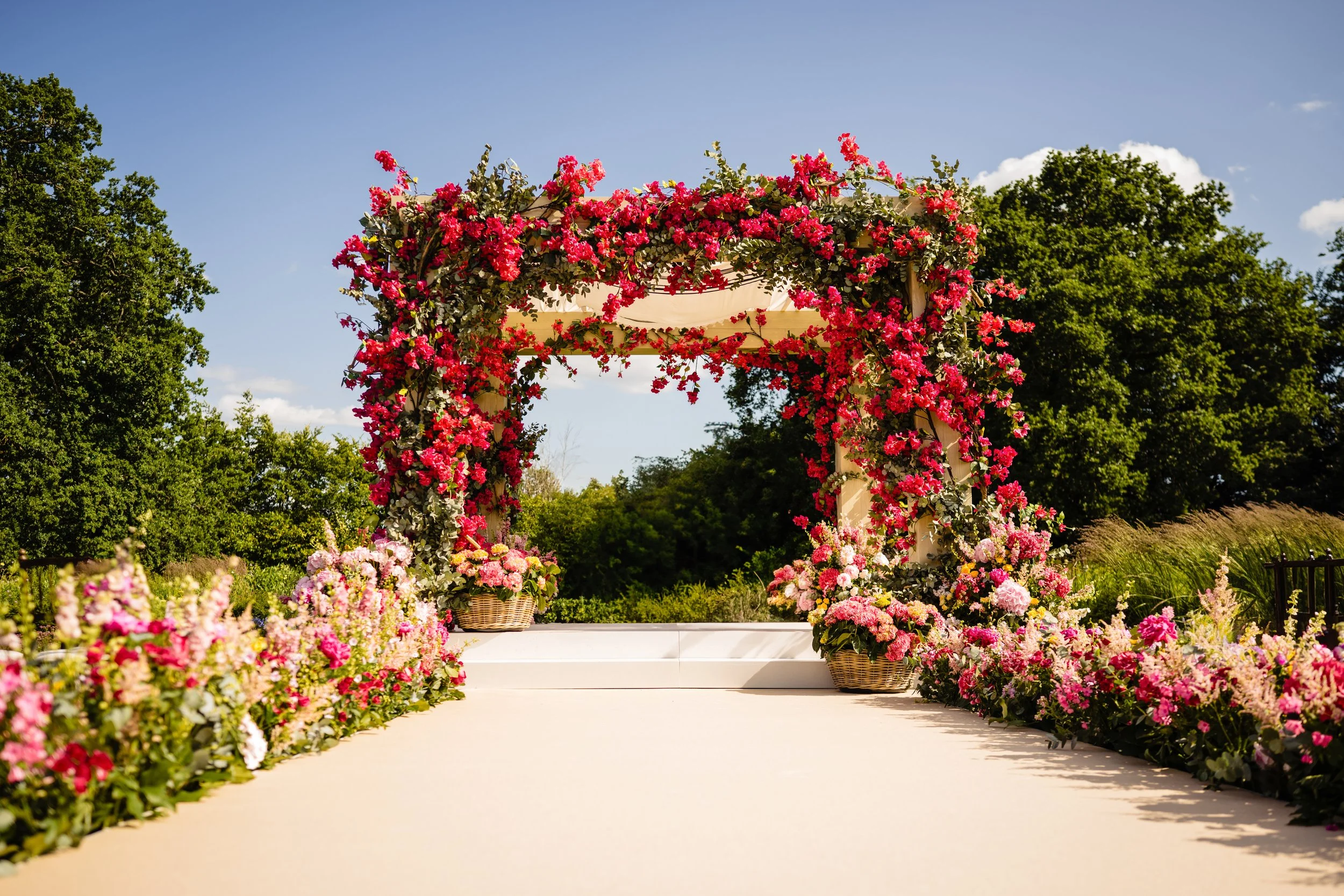 A wedding ceremony setup outdoors with a floral archway covered in pink and red flowers, surrounded by more flowers along the aisle, with green trees and a blue sky in the background.