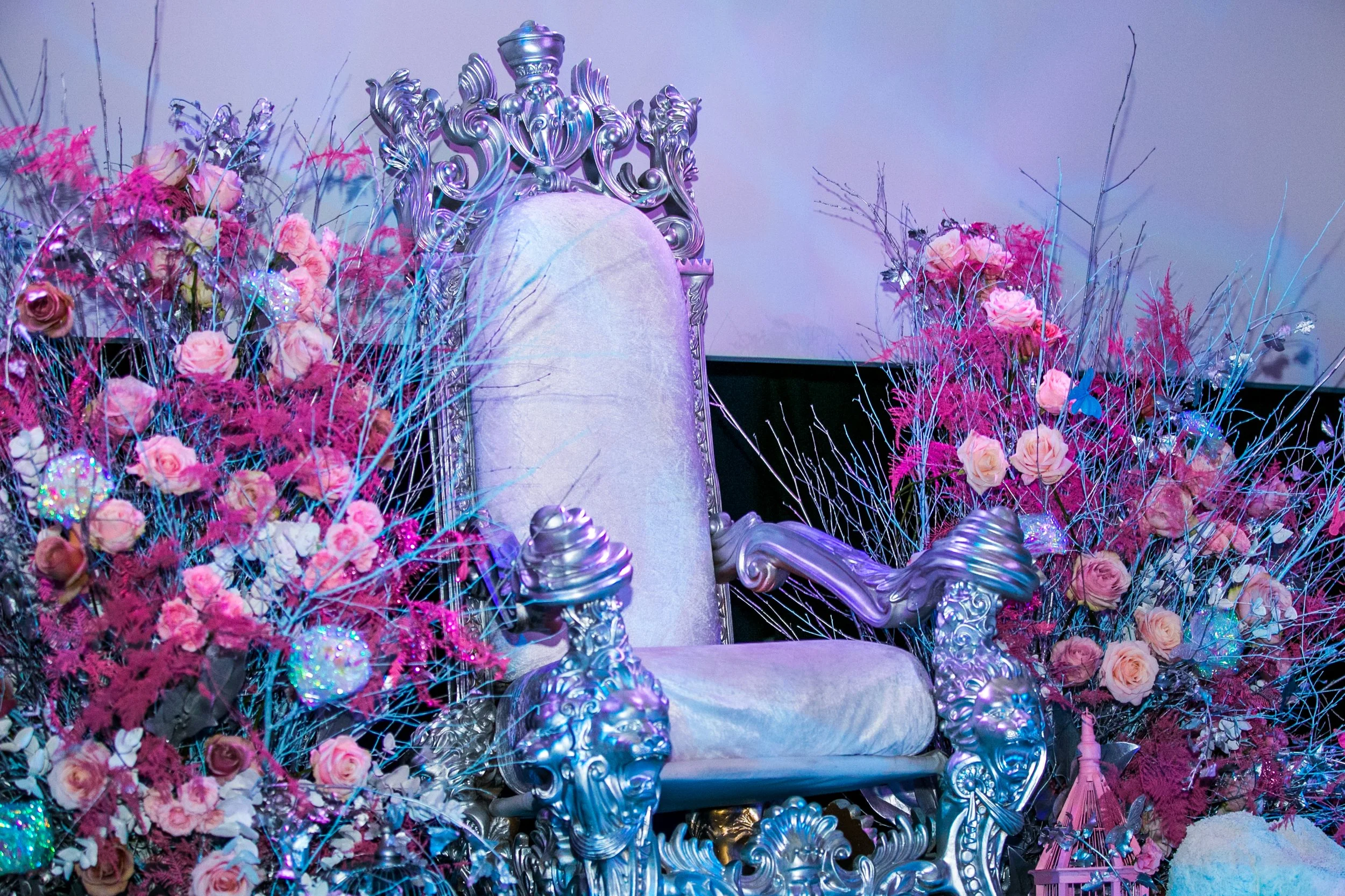 A silver ornate throne with floral decorations and pink roses, illuminated by colorful lights. ICE PALACE BAT MITZVAH at Old Billingsgate 