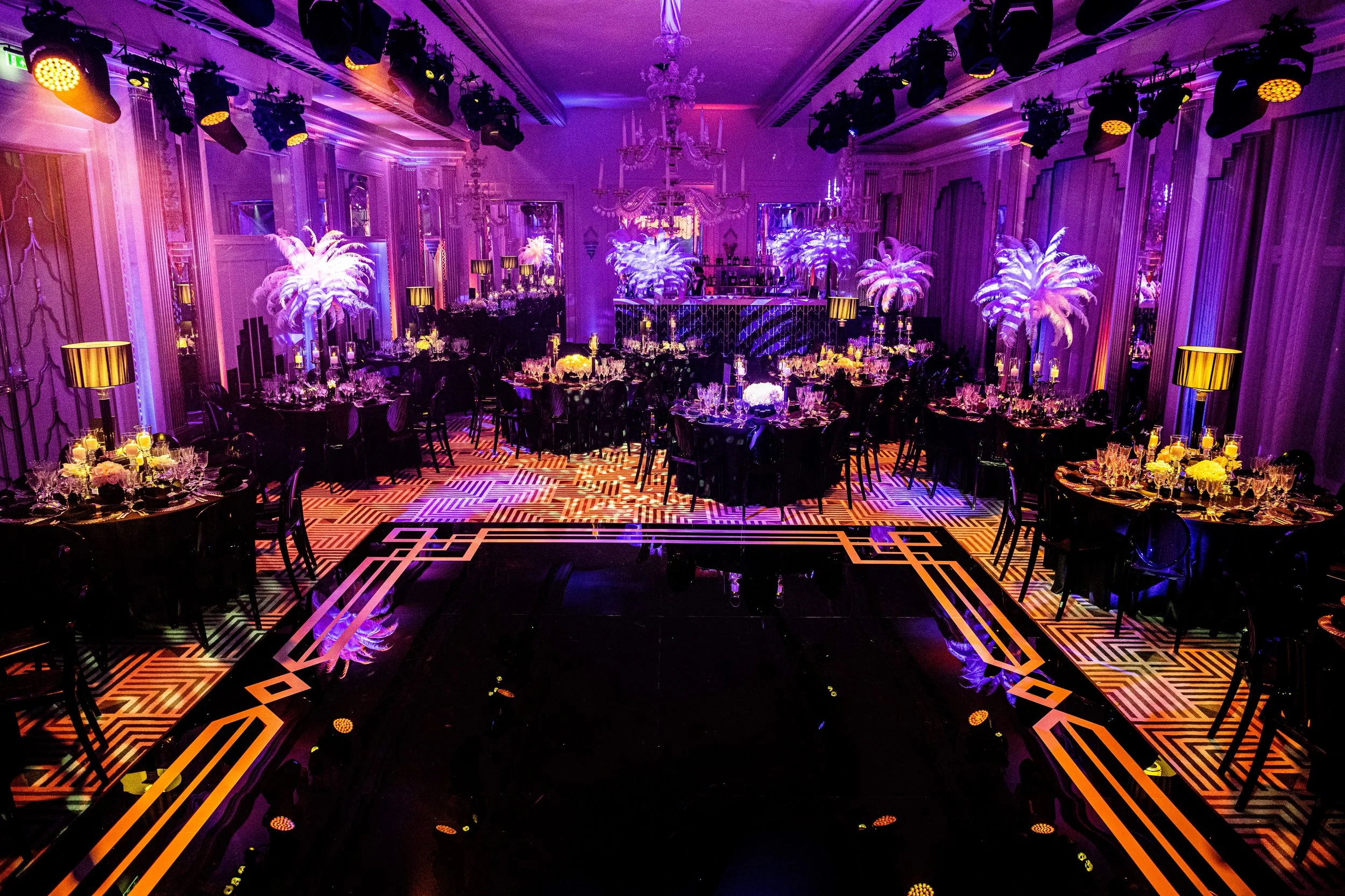 Claridge's decorated in a 50's themed party with purple lighting, tall feathered palm centerpieces, chandeliers, and arranged dinner tables with candle centerpieces.