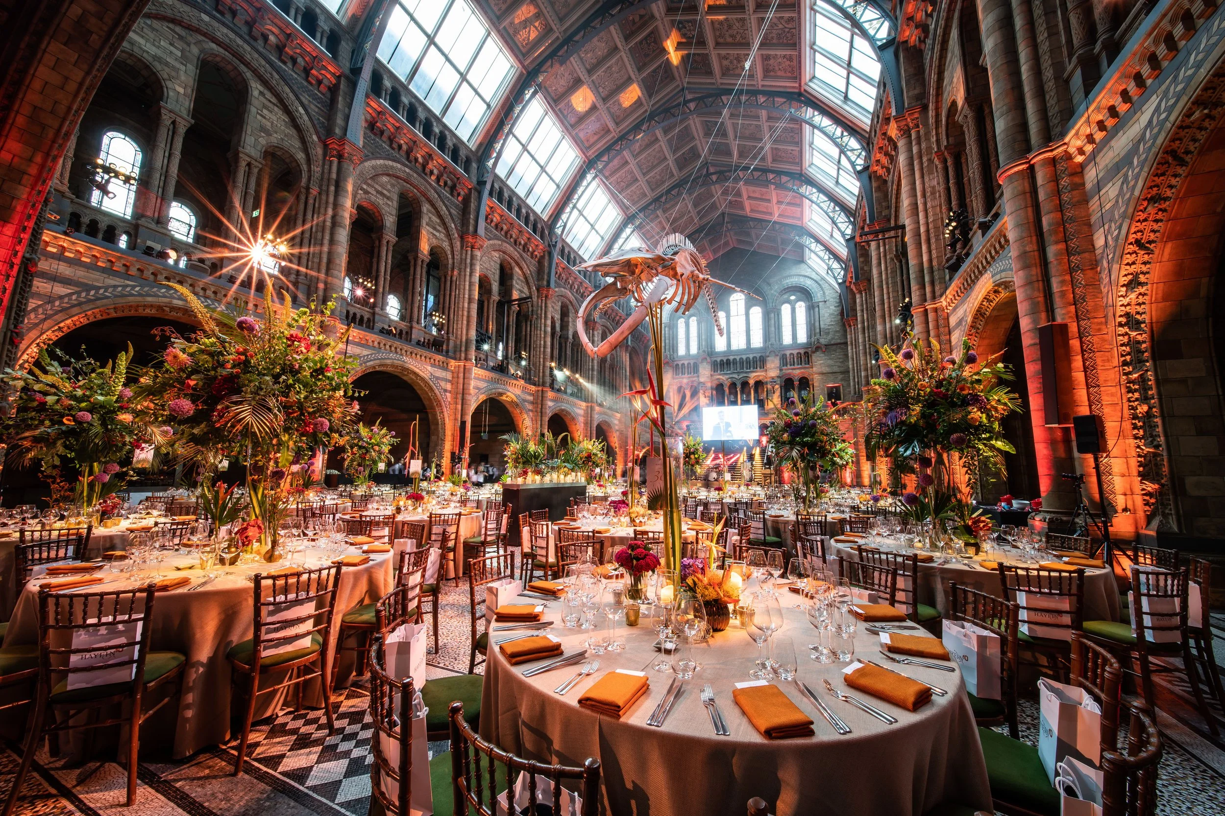 Gourmet banquet setup inside a large, ornate historic building with high arched glass ceilings, floral centerpieces, and elegant table settings at The Natural History Museum