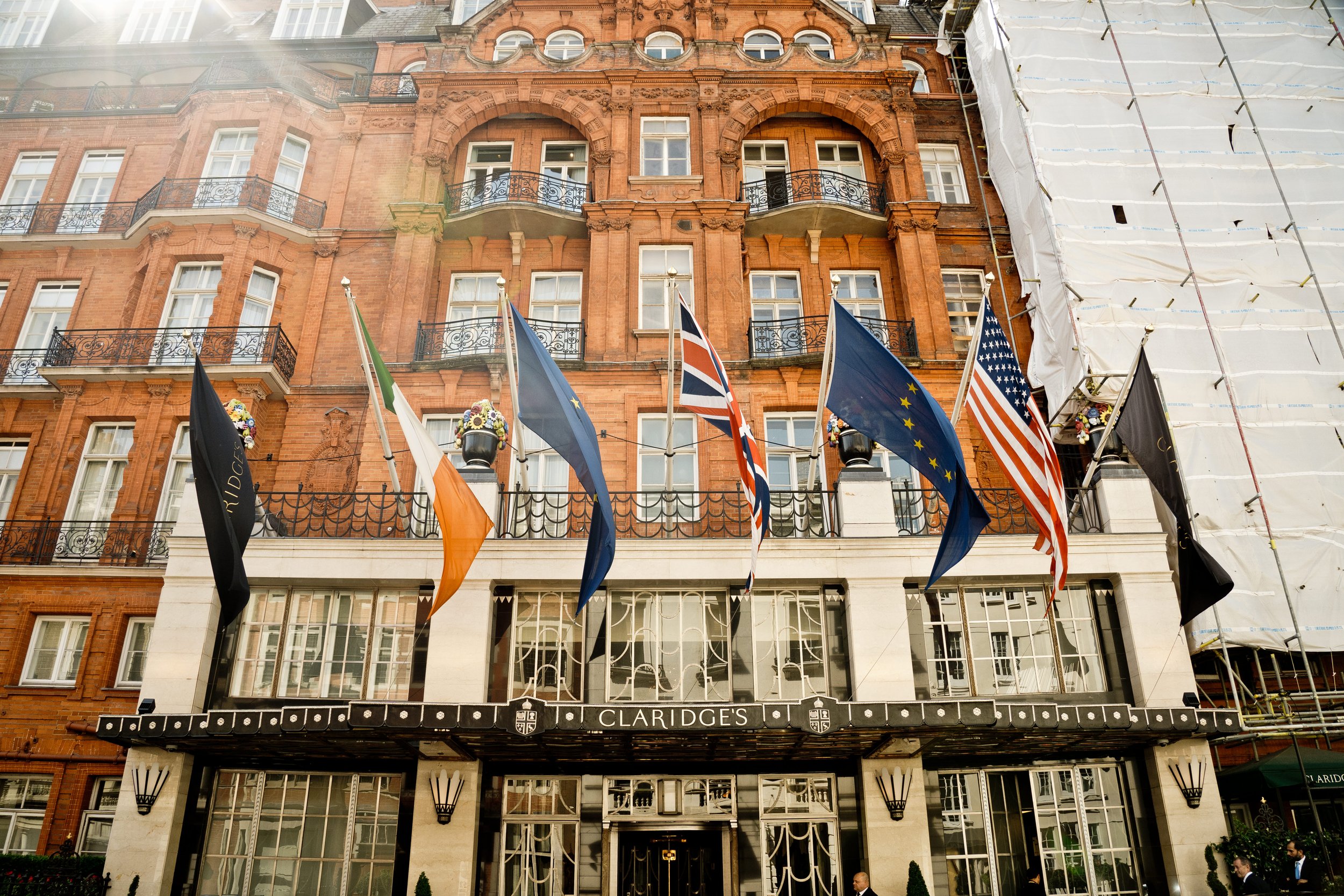 The facade of a brick building with multiple floors, ornate architectural details, balconies, and windows. Flags are displayed in front of the building, and a sign reads "CLARIDGES" at the entrance. Part of the building is covered with white scaffold