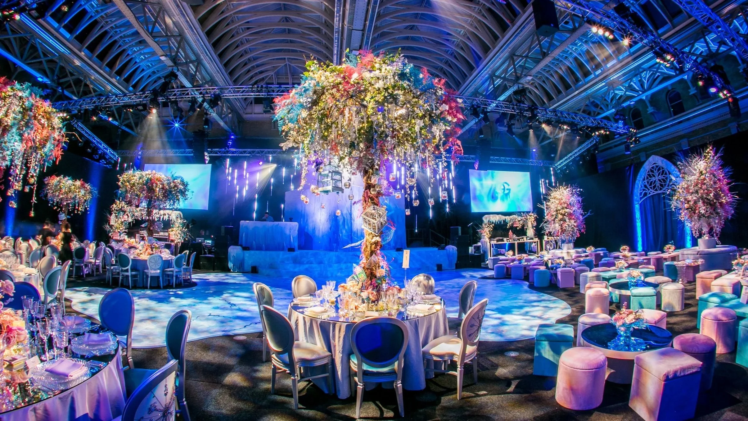 Bat Mitzvah celebration at the Old Billingsgate event hall with circular tables, elegant chairs, and pastel-colored stools, illuminated by blue lighting and adorned with large floral centerpieces hanging from the ceiling for a Ice Palace look.