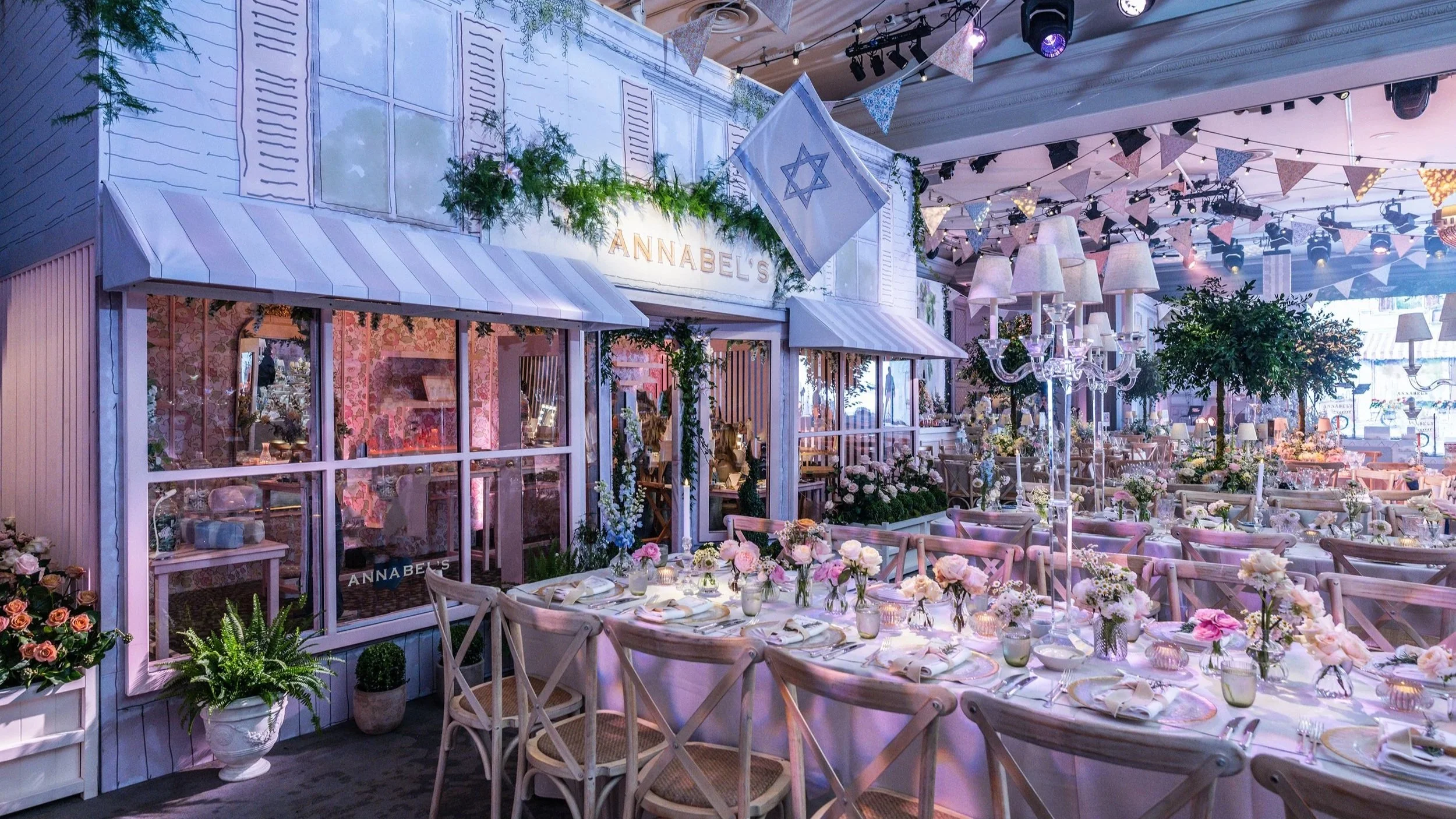 The Marriot JW Grosvenor House decorated with pink and white flowers, string lights, and bunting banners. There are tables set with plates, glasses, and floral centerpieces. Hamptons inspired design with an Israeli flag hangs in the background