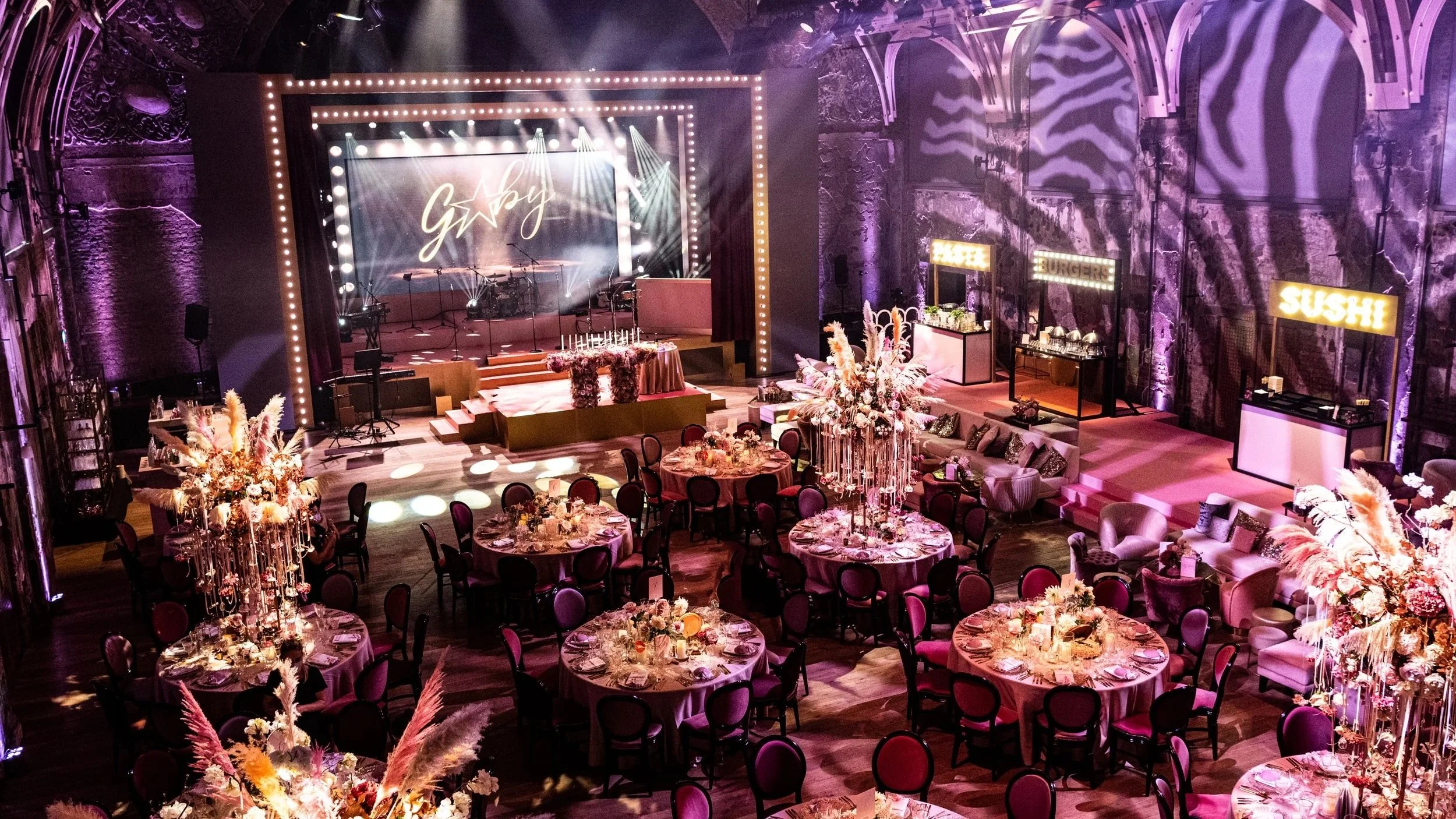 Battersea Arts Centre decorated with floral centerpieces, a stage with musical instruments for hollywood glamour theme.  Pastel and purple lighting highlights the ornate architecture, with signs for burger and sushi on the sides.