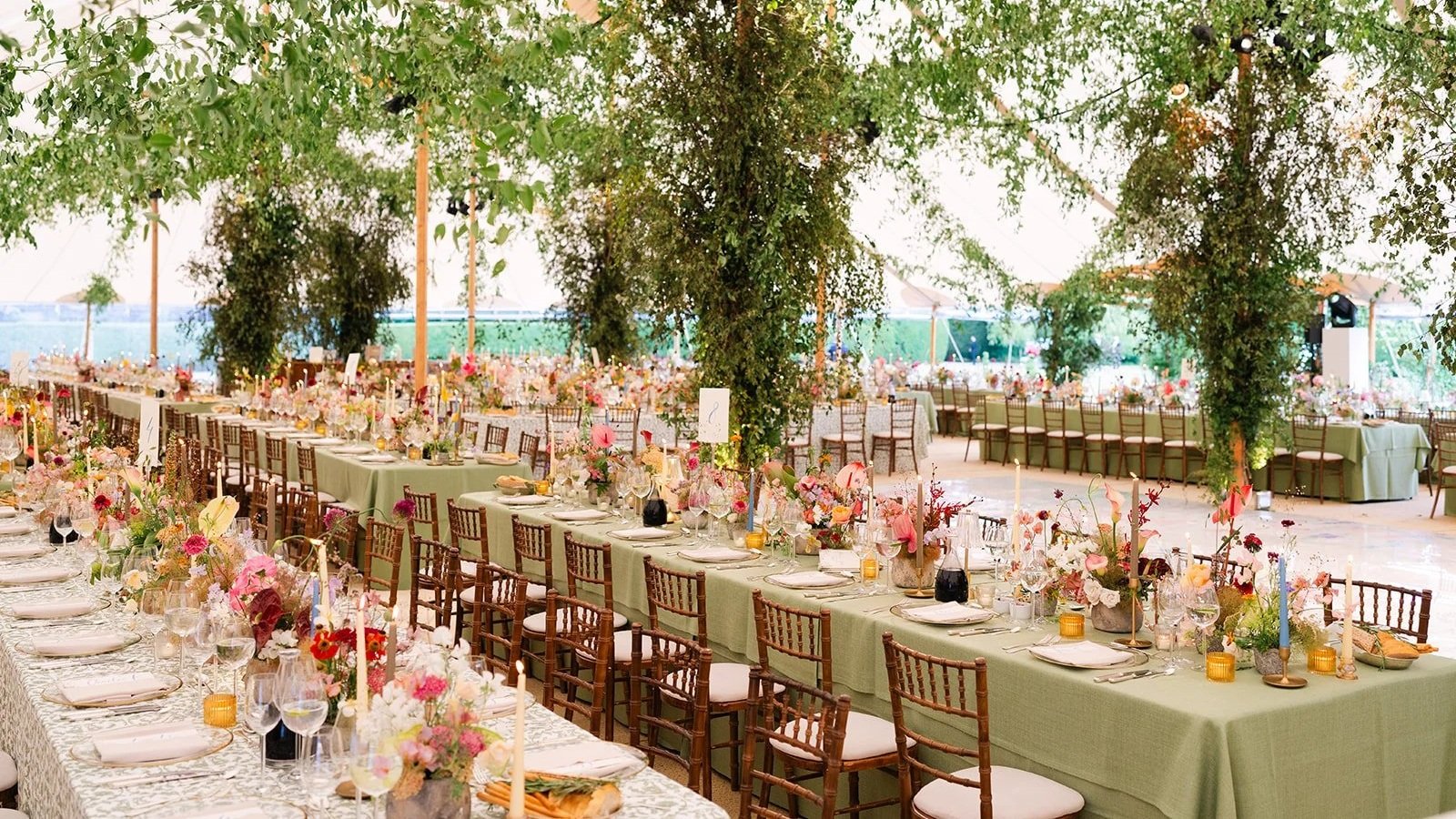 Private Residents Weddings in Hertfordshire  long tables covered in green tablecloths, adorned with pink, white, and peach flowers, candles, glassware, and tableware, under a tent with hanging greenery and trees.
