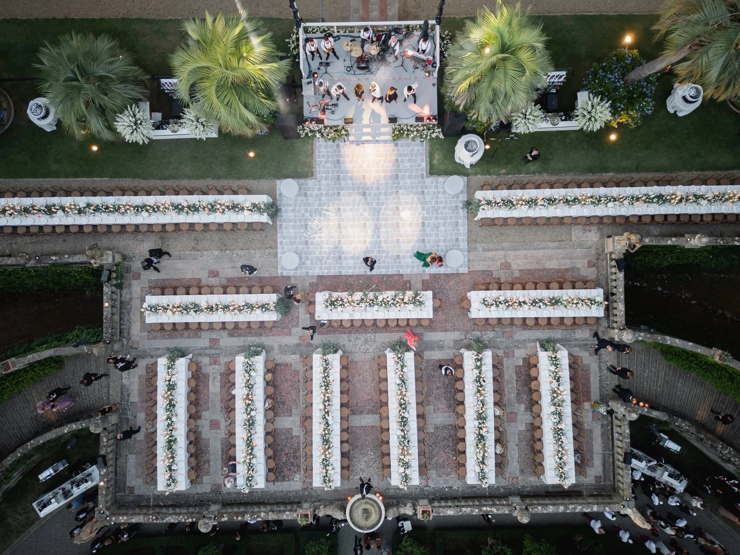 An aerial view of an outdoor wedding reception setup with tables decorated with white and pink flowers, palm trees, and a band playing on a small stage, with guests walking around at Villa Palmieri in Florence