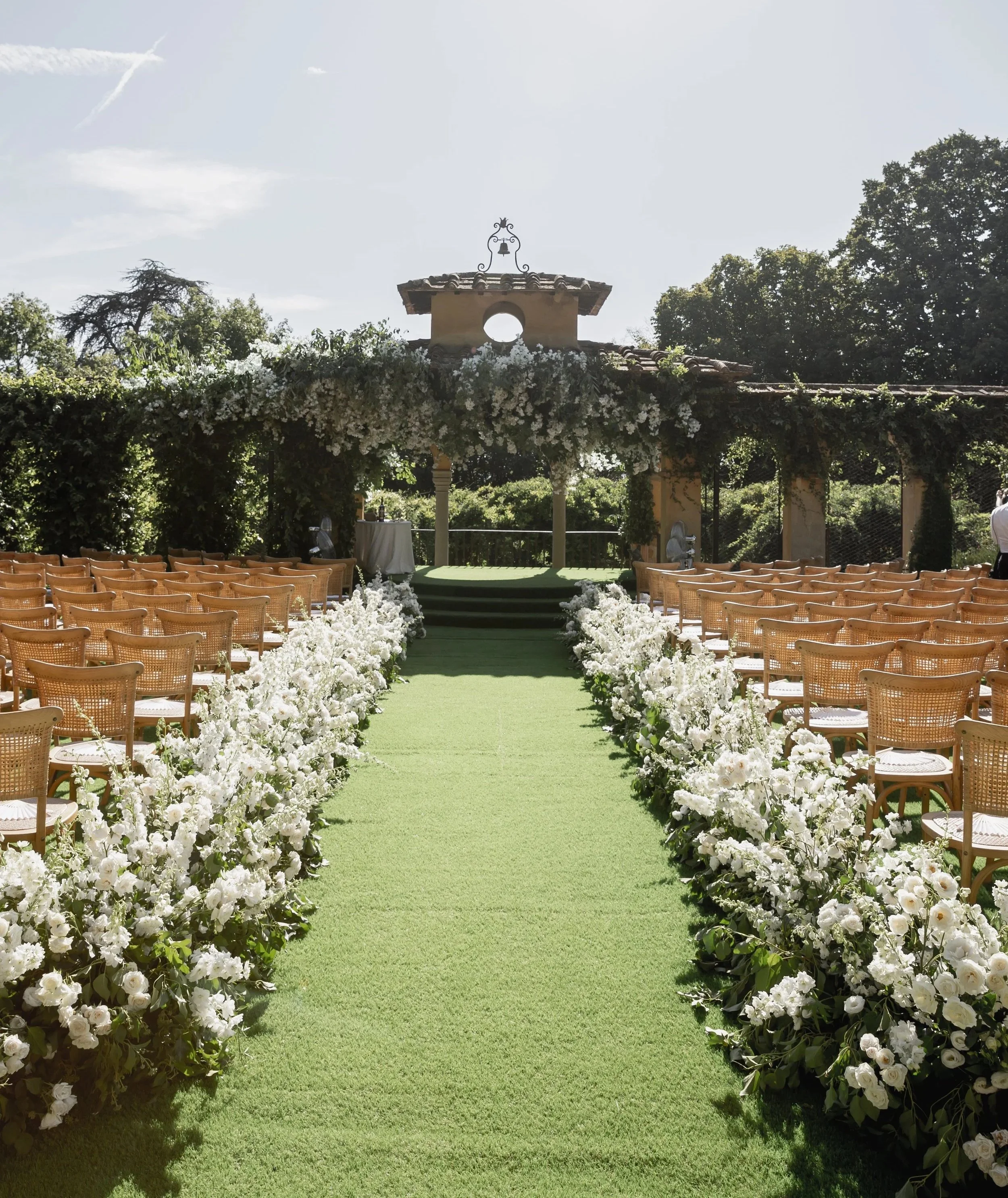 Outdoor wedding ceremony at Villa Palmieri with rows of brown chairs on either side of a green aisle, decorated with white flowers, leading to a decorated altar with white flowers and a small building with a bell on top in the background.