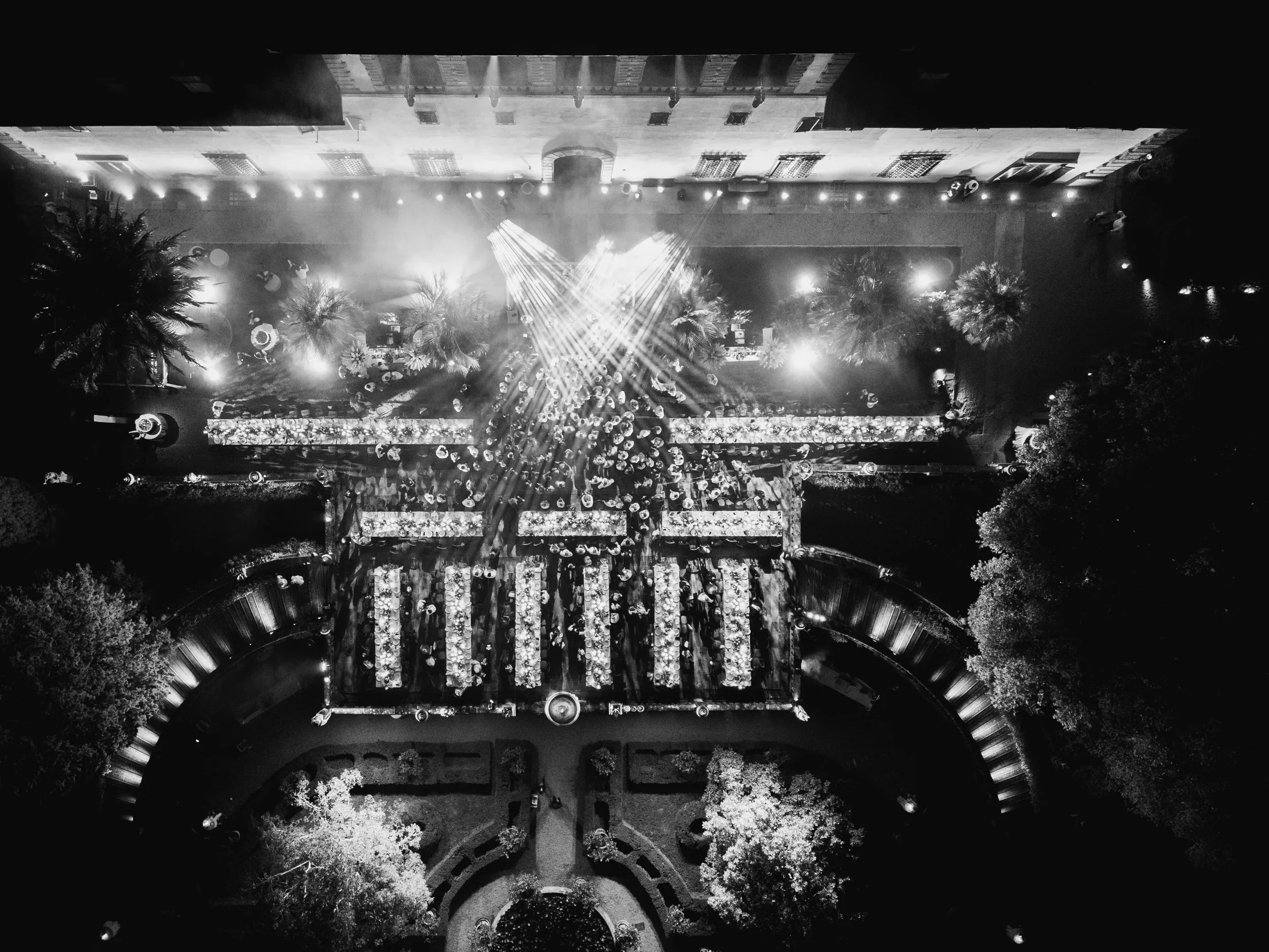 A black and white aerial view of a decorated outdoor event space with a stage, tables, and trees illuminated by lights at Villa Palmieri in Florence