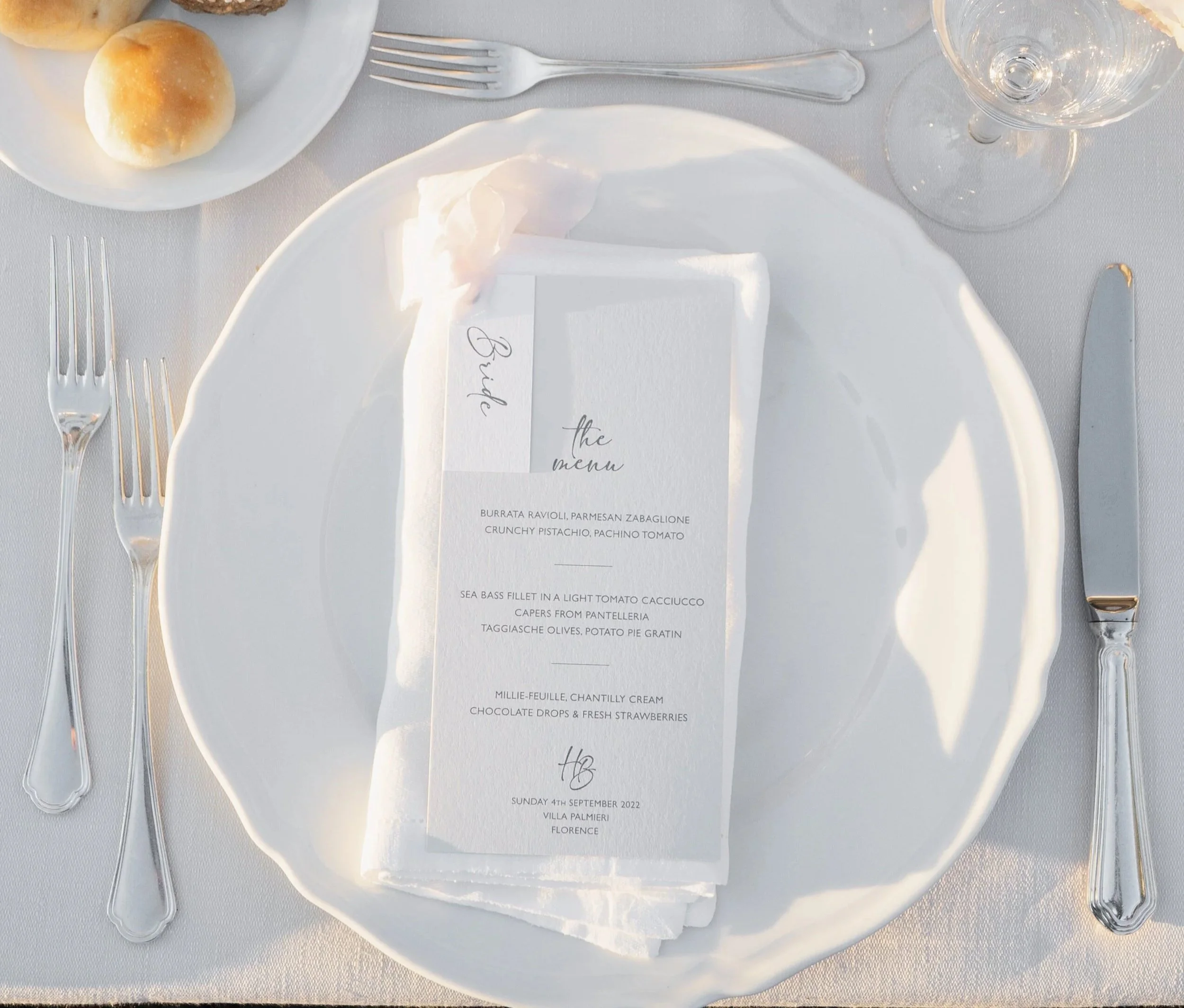 Close-up of a formal dining table setting with white plates, silver cutlery, a wine glass, and a printed menu on a white napkin. The menu lists dishes and has a handwritten date and location at the bottom.