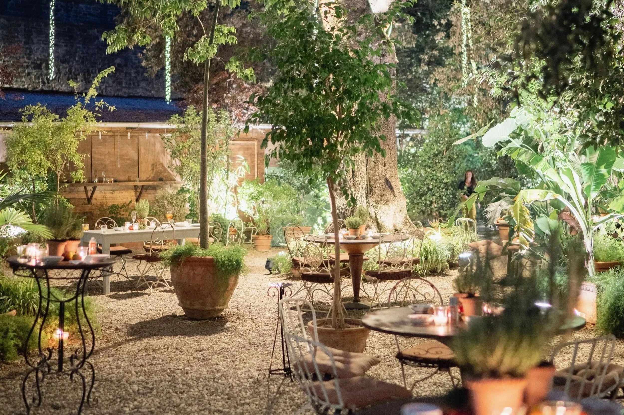 An outdoor dining area at night with round tables and metal chairs, surrounded by lush green plants and large potted trees, with a wooden shed and string lights in the background  in Tuscany at Serre Torrigiani Bistrot