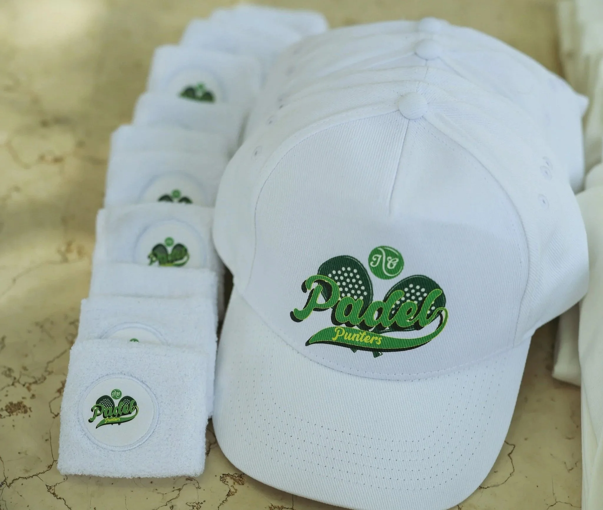 Line of white baseball caps with green and black logo reading Padel on the front, laid on a beige cracked surface at Anantara Villa Padierna Resort in Marbella