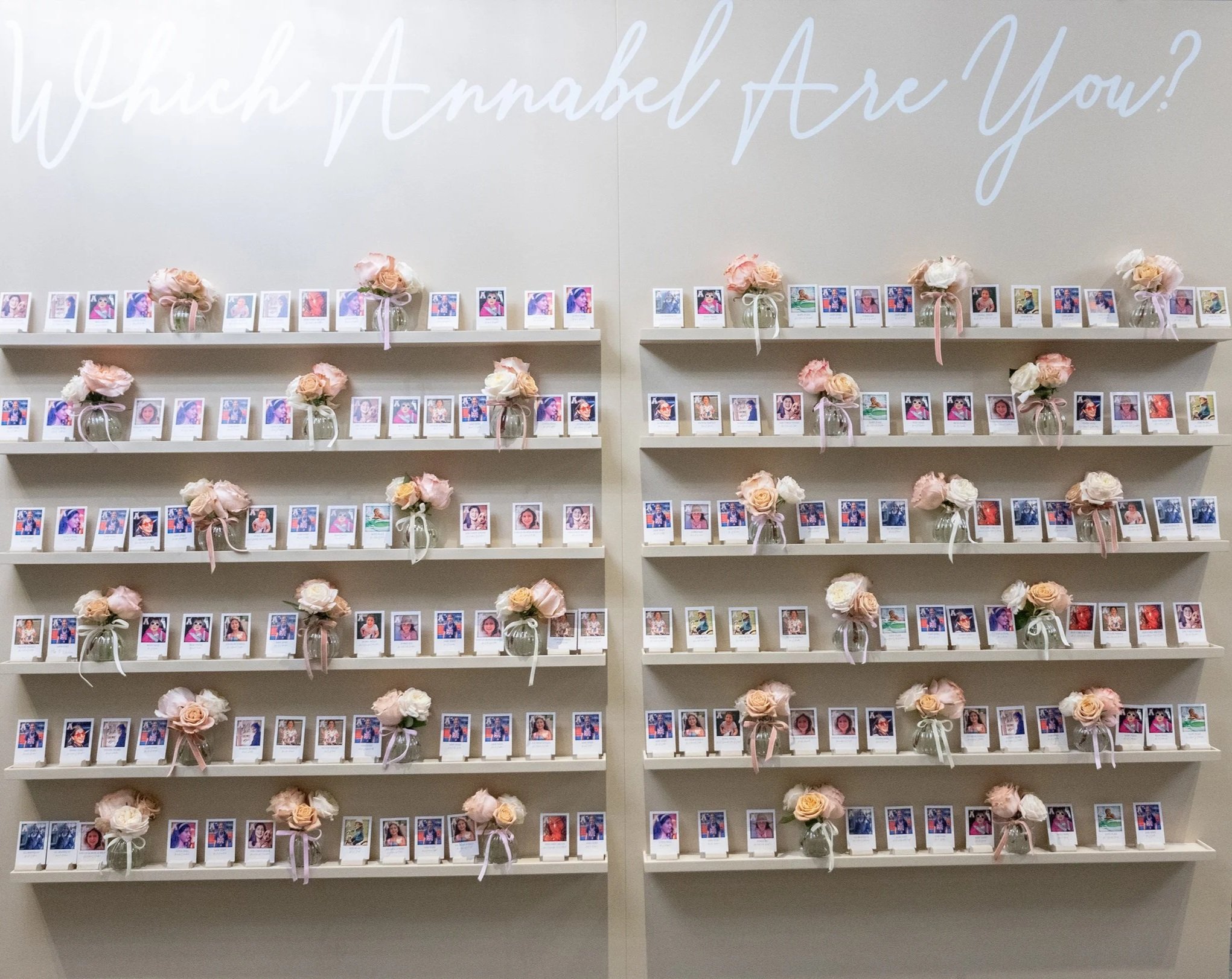Display of small photographs of children and adults, arranged on white shelves with pink and white floral bouquets in glass vases, against a beige wall with large white cursive text that reads 'Happily Ever After!'