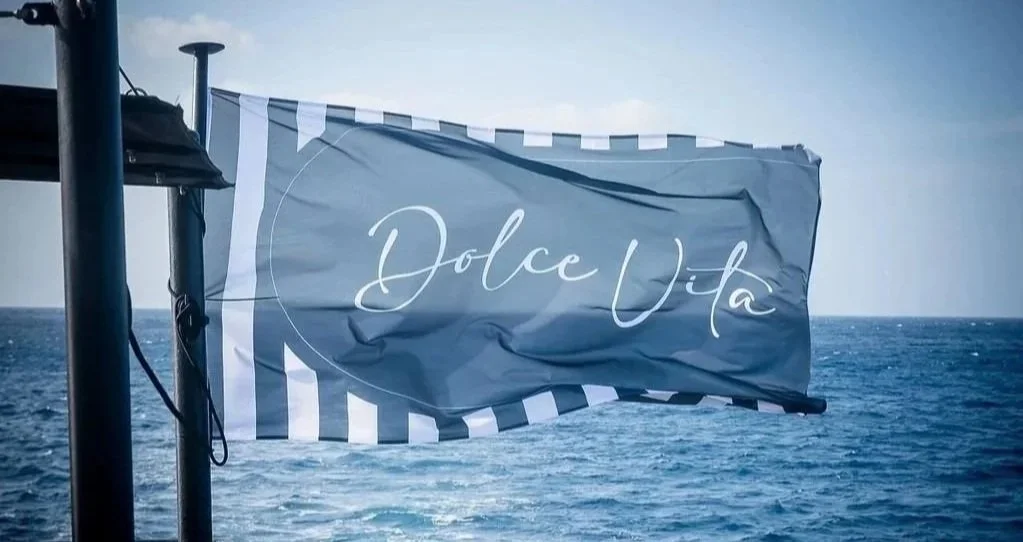 A flag with the text 'Dolce Vita' on it, hanging on a pole over the ocean under a cloudy sky.