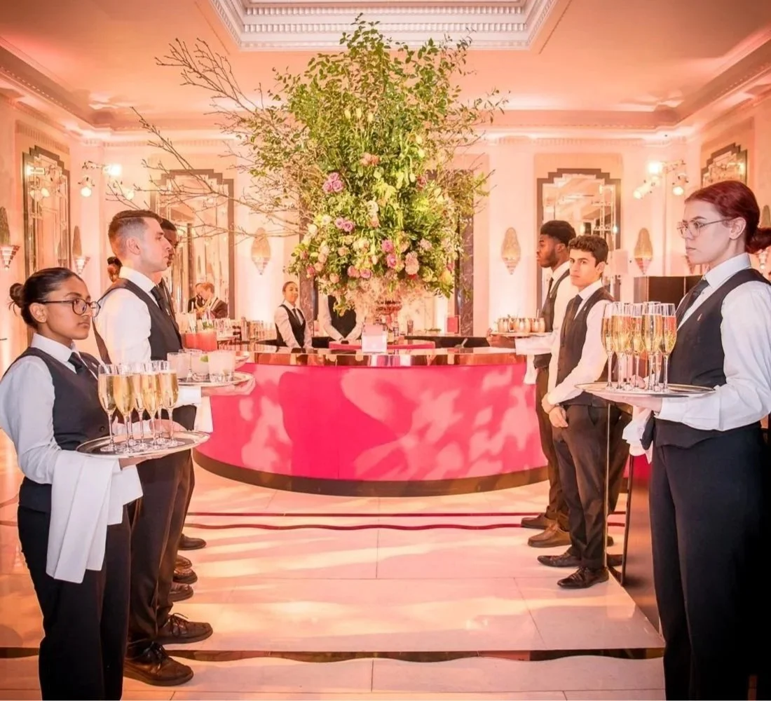 Waitstaff standing in a line at a luxurious event, each holding a tray with glasses of champagne, facing a large floral arrangement at a pink bar in an elegant, well-lit room with soft lighting and ornate decor.