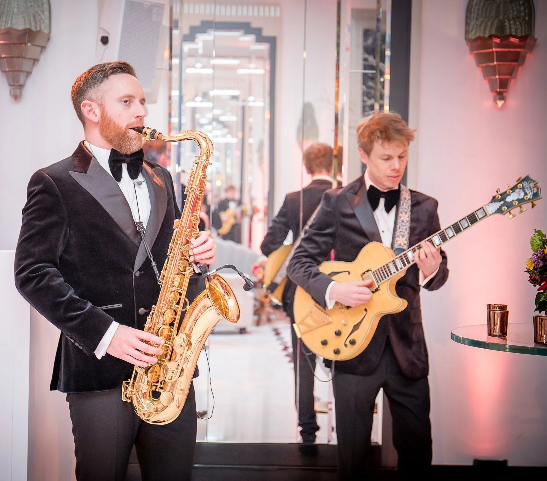 Two musicians in tuxedos performing indoors, one playing a gold saxophone and the other a light-colored electric guitar, with modern decor and wall sconces.