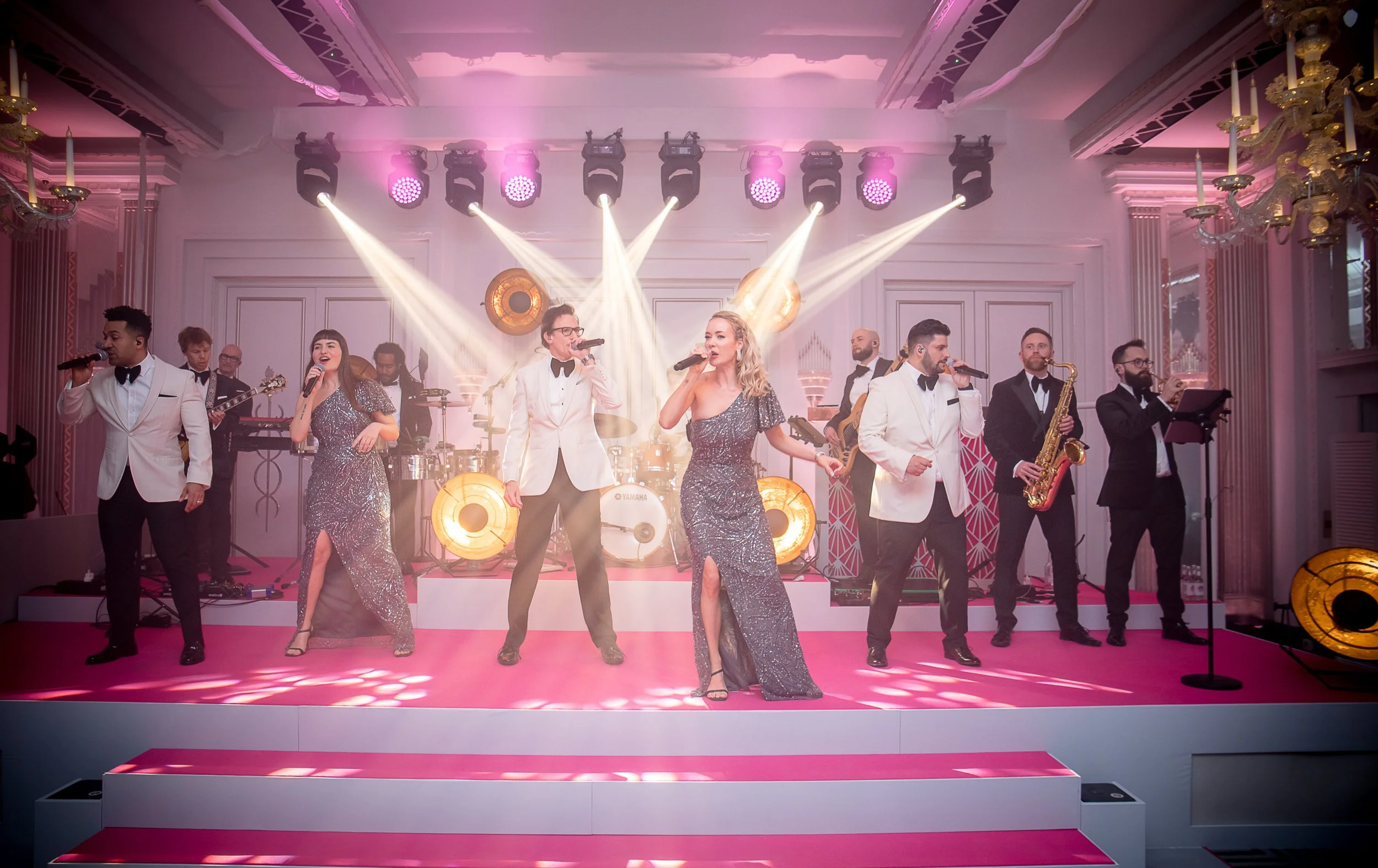 A band performing on stage in formal attire, with two female singers in sequined dresses at the center, surrounded by male musicians and singers, with stage lighting and elegant decor, in a grand venue.