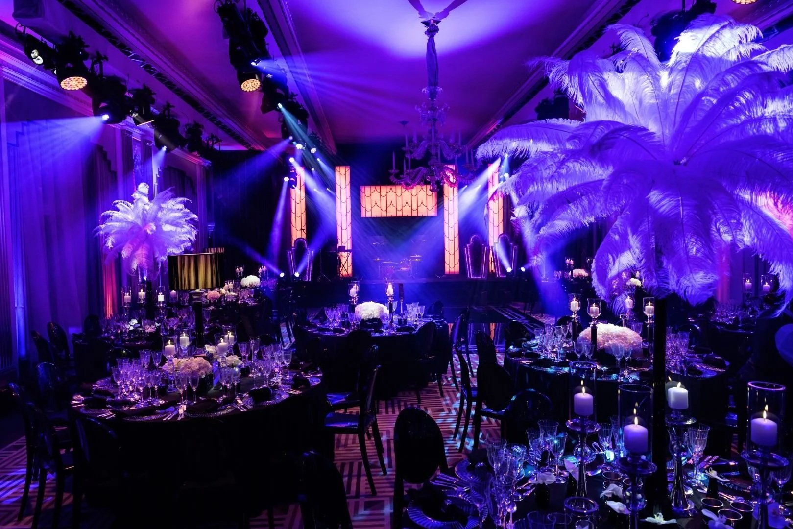 A lavish event space decorated with purple lighting, large white feathered palm trees, and elegant table settings with candles, flowers, and glassware, featuring a stage with vertical light panels and a chandelier hanging from the ceiling.