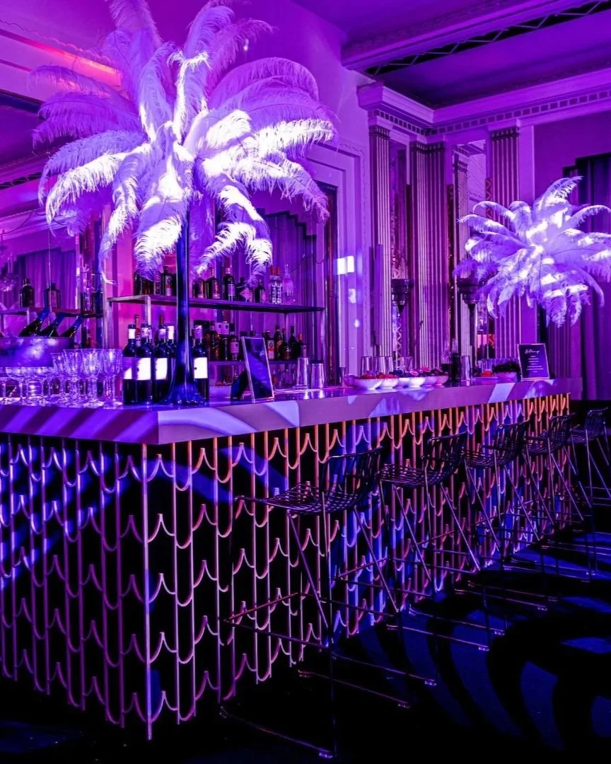 Decorative bar with purple lighting, palm tree-shaped centerpieces with illuminated white fronds, bottles of alcohol on shelves, glasses on the bar counter, and modern bar stools. Claridge's decorated in a 50's themed party 