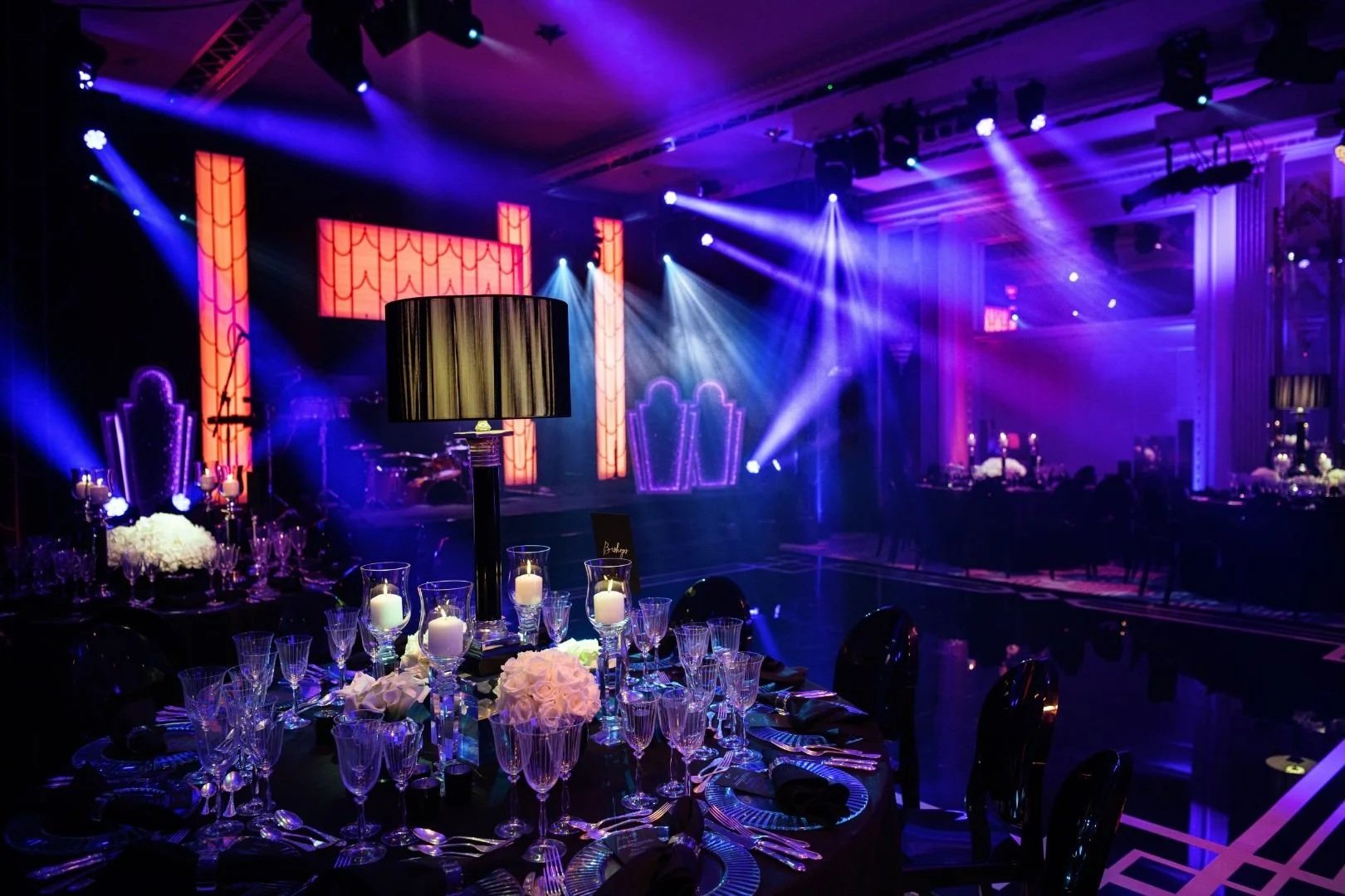 Elegant event space with tables set with candles, flowers, and glassware, illuminated by purple and blue stage lights, featuring a stage with decorative columns and chairs in the background.