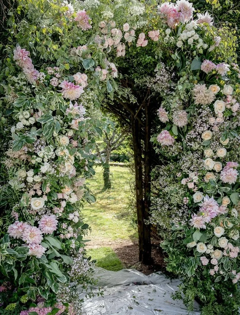 A floral archway decorated with pink and white roses, dahlias, and greenery, leading outdoors to a garden or park area.