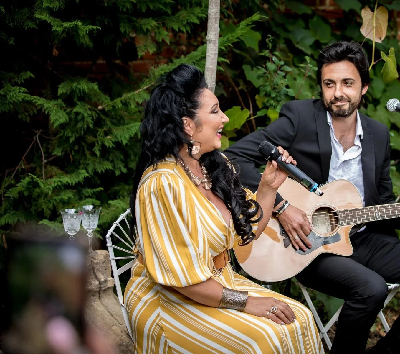 A woman in a yellow striped dress singing into a microphone while sitting, next to a man in a suit playing an acoustic guitar, outdoors surrounded by green foliage.