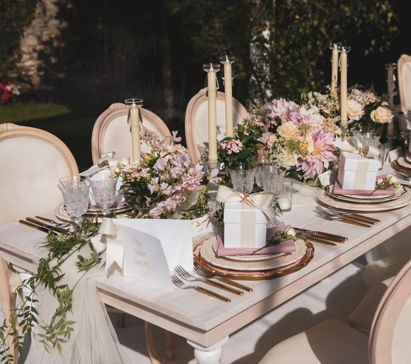 Elegant outdoor wedding or event table setting with floral centerpieces, candles, gift boxes, and fine china.