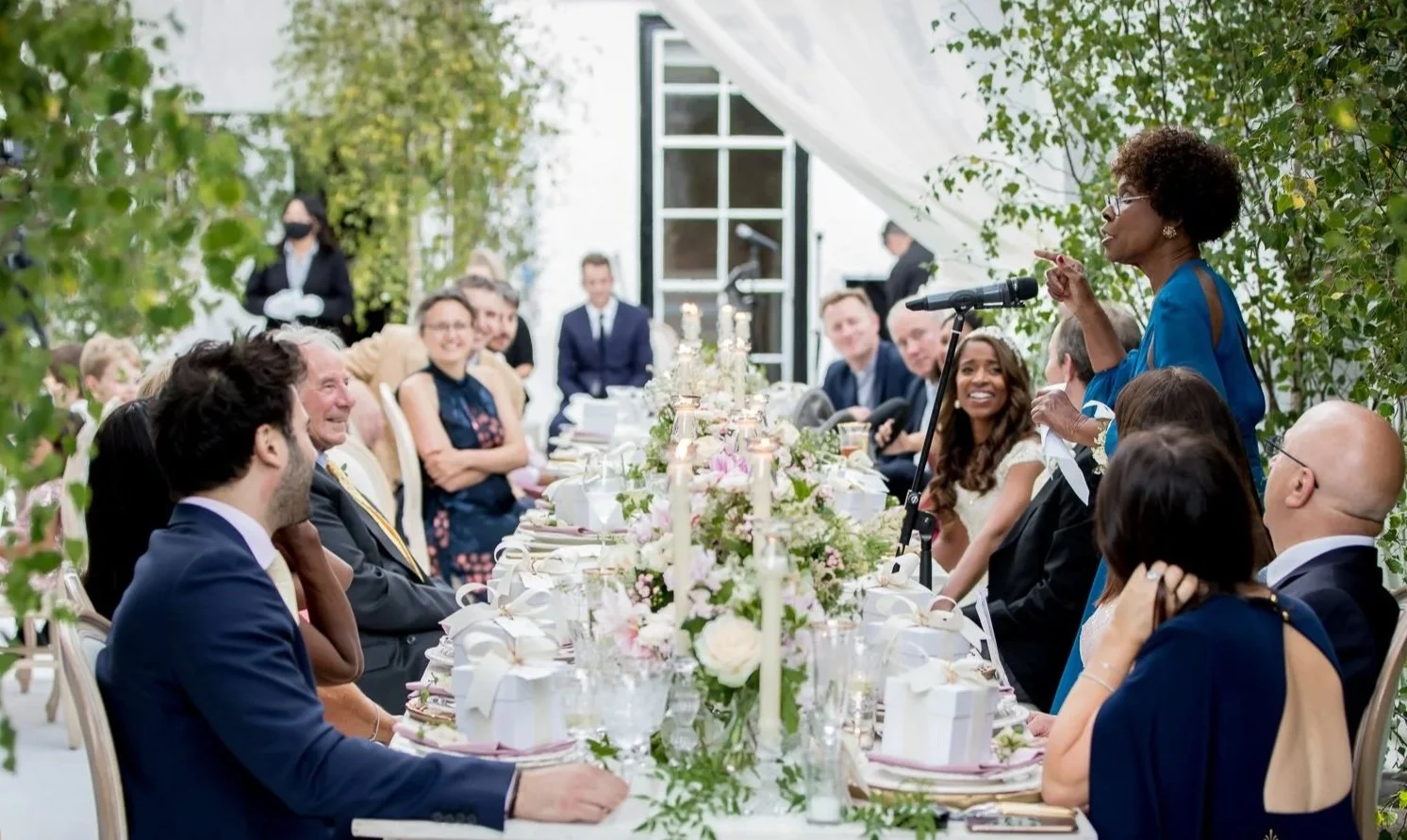 A woman giving a speech at a formal outdoor event, with guests seated at a decorated table, surrounded by greenery.