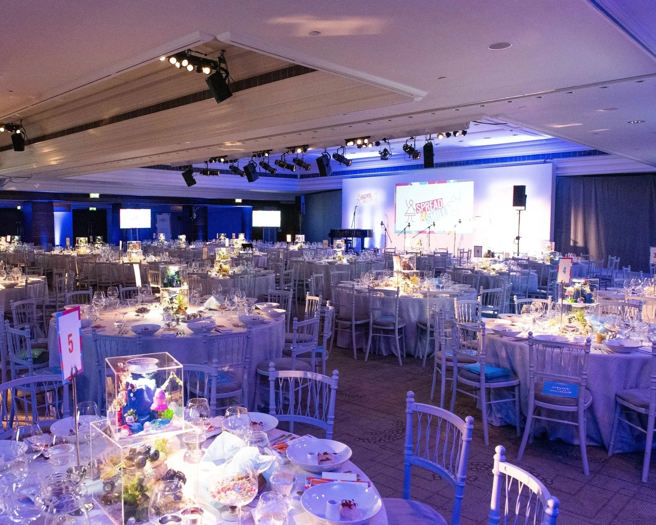 Event venue decorated with round tables, centerpieces, and a stage with large screens displaying the logo 'Spread a Smile.'