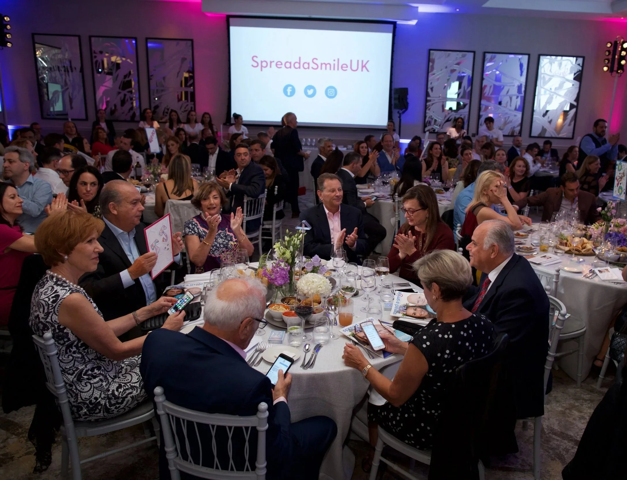A formal event with many people sitting at round tables, some clapping, with flowers and food on the tables. There is a large screen displaying "SpredaSmileUK" with social media icons, and the room is decorated with purple and blue lighting.
