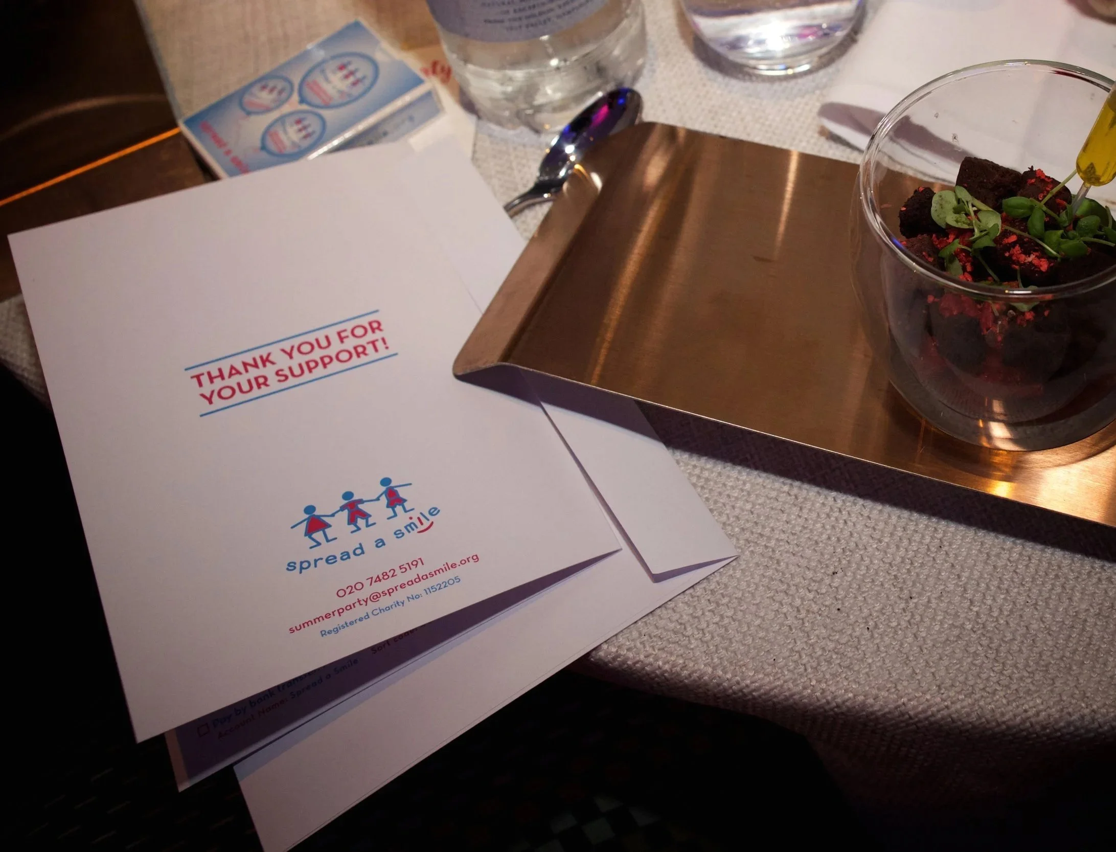 A close-up of a table setting with a white paper with red and blue text that reads 'Thank you for your support,' a clear plastic cup with a colorful plant inside, a glass of water, and some small cards or flyers. The items are placed on a table with 