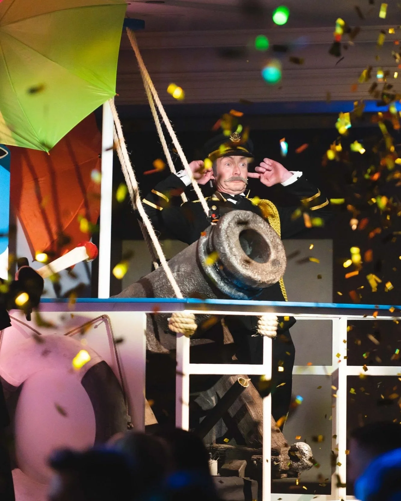 A person dressed as a ship captain on a decorated boat with a green umbrella and a cannon, with colorful confetti falling around and in front of the person.