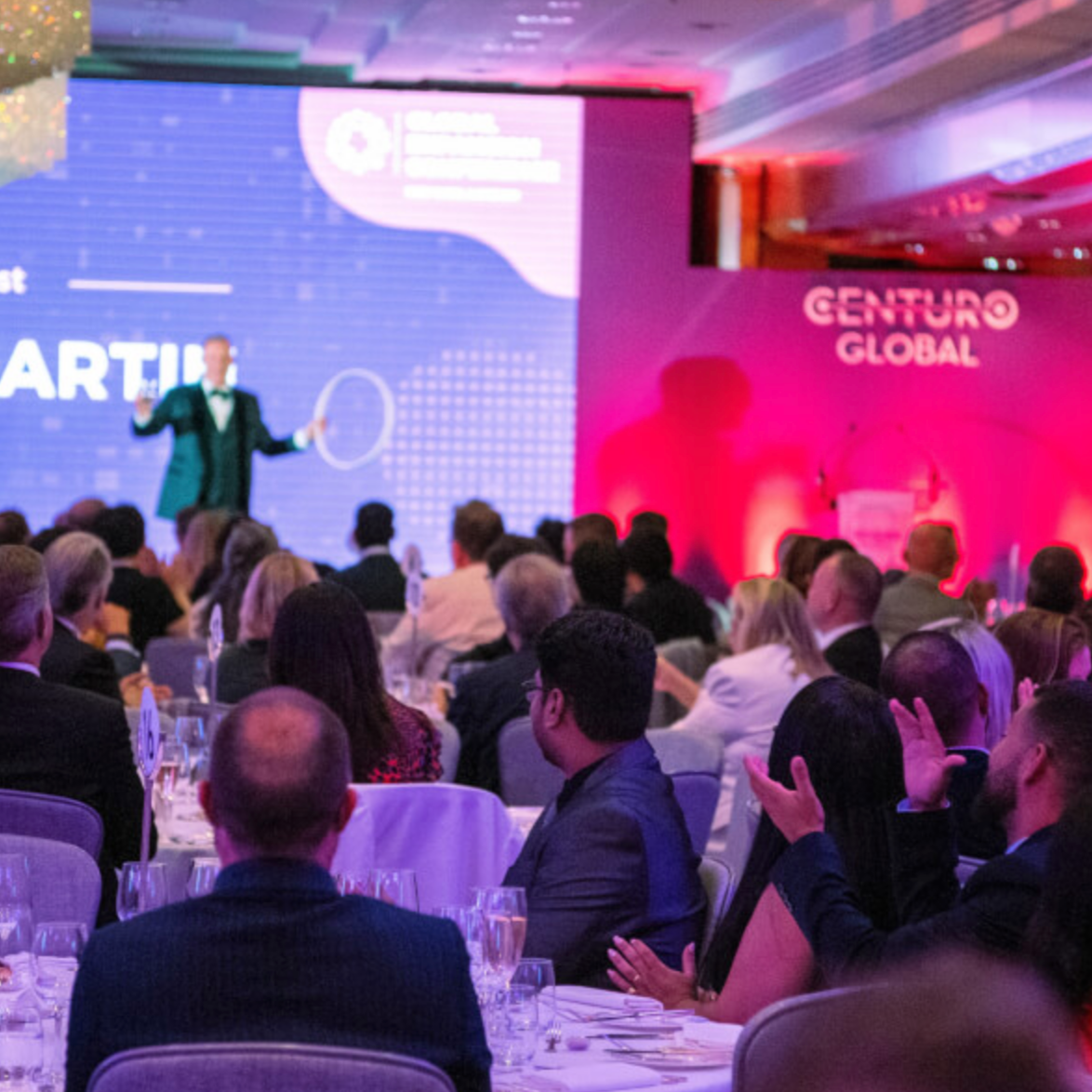 A formal event with an audience seated at round tables, facing a stage with a speaker presenting. The stage has a large screen displaying graphics and text, and the wall behind the stage has the words 'CENTURO GLOBAL' illuminated in pink. The audienc