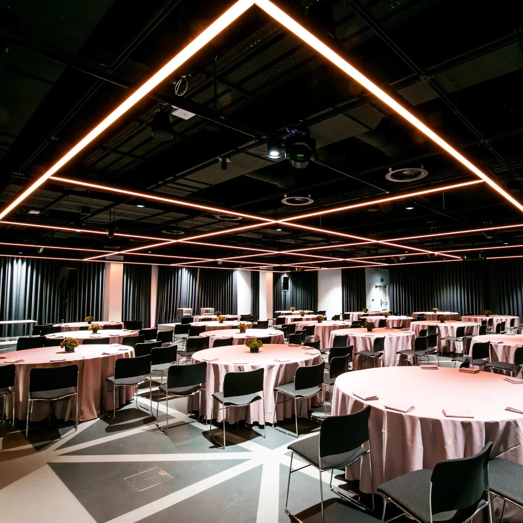 A large conference room with round tables covered in pink tablecloths, each decorated with small flower arrangements. The room has black chairs, and the ceiling features modern lighting fixtures with rectangular light strips forming a grid pattern.